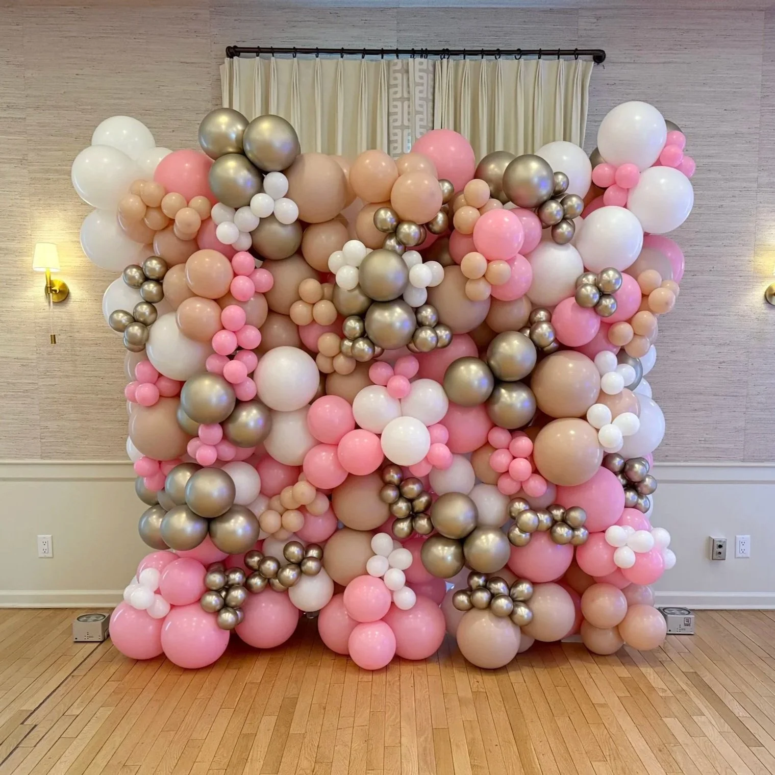 Decorative balloon wall with pink, white, tan, and metallic gold balloons arranged in clusters, set against a wall with curtains, light fixtures, and wood flooring.