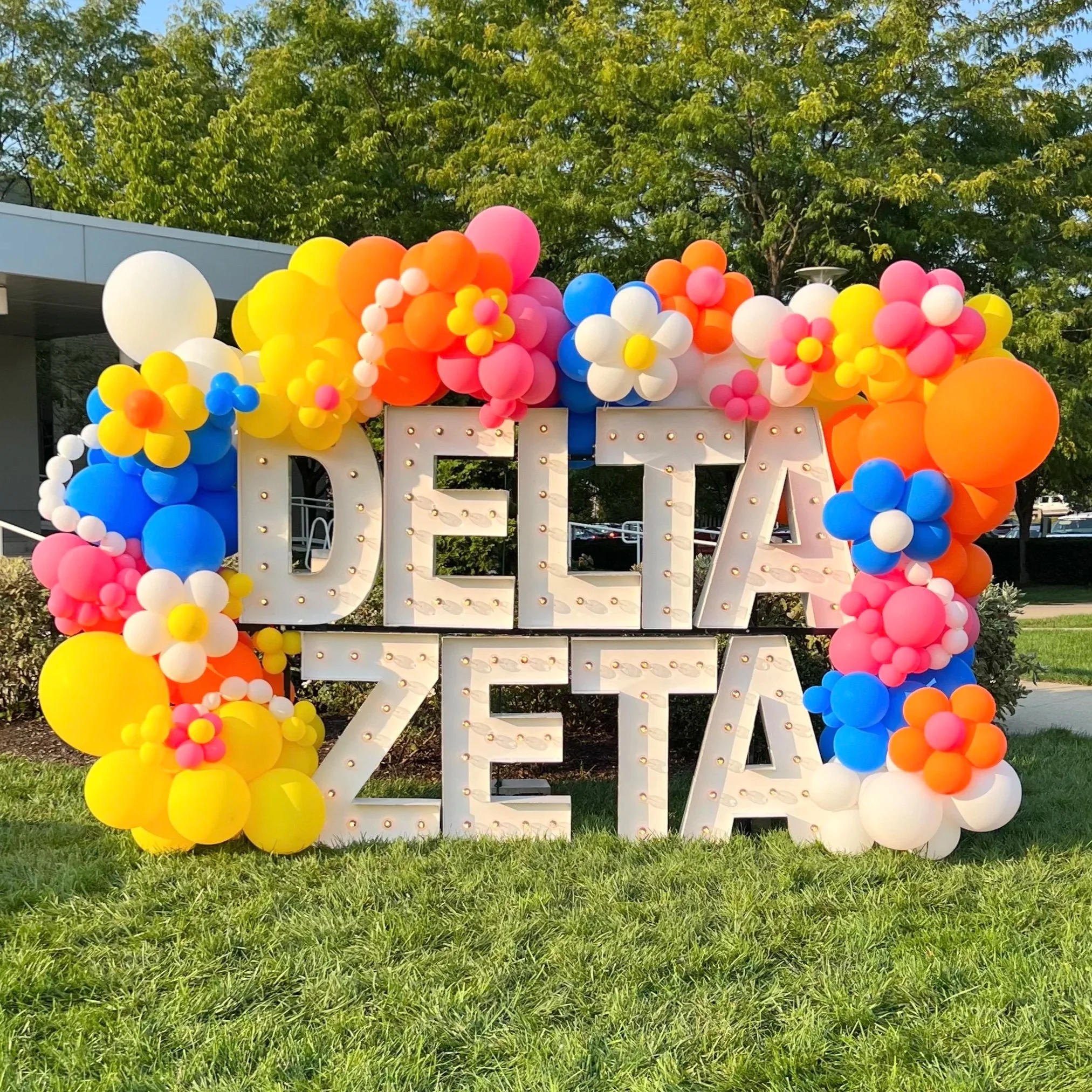 Decorative sign with the word 'DEJA' in large letters, surrounded by colorful balloons shaped like flowers, in shades of yellow, pink, white, blue, and orange, set outdoors on a grassy area with trees in the background.