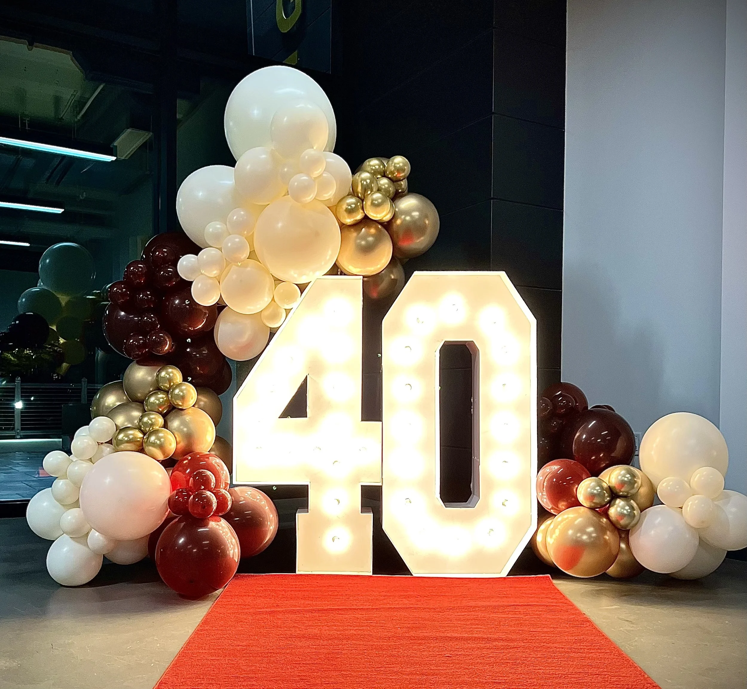 Large illuminated number 40 surrounded by an arrangement of balloons in white, gold, red, and dark colors, on a red carpet, celebrating a 40th birthday.
