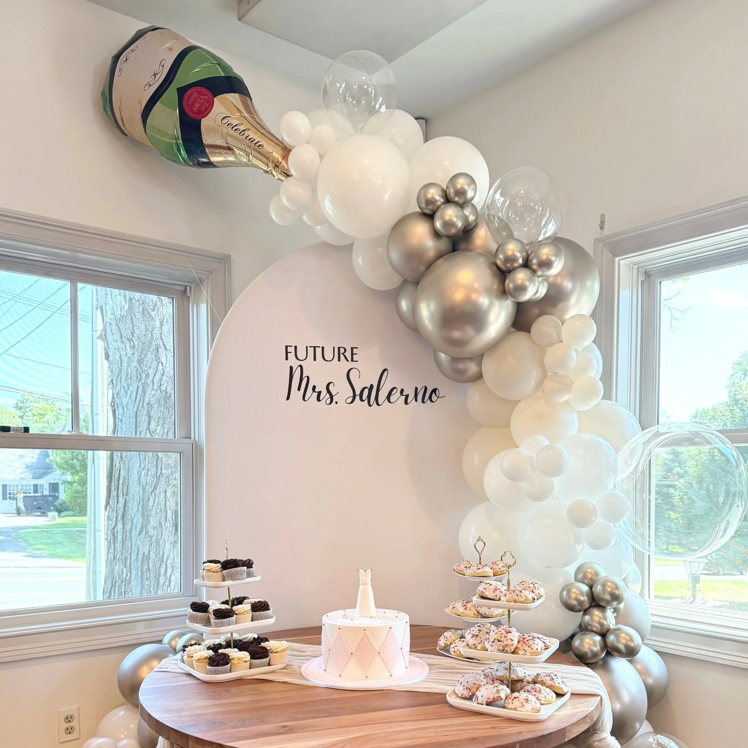 Celebration with a balloon arch in silver, white, and clear balloons, a cake on a wooden table, cupcakes, and donuts, with a sign reading "Future Mrs. Salerno."
