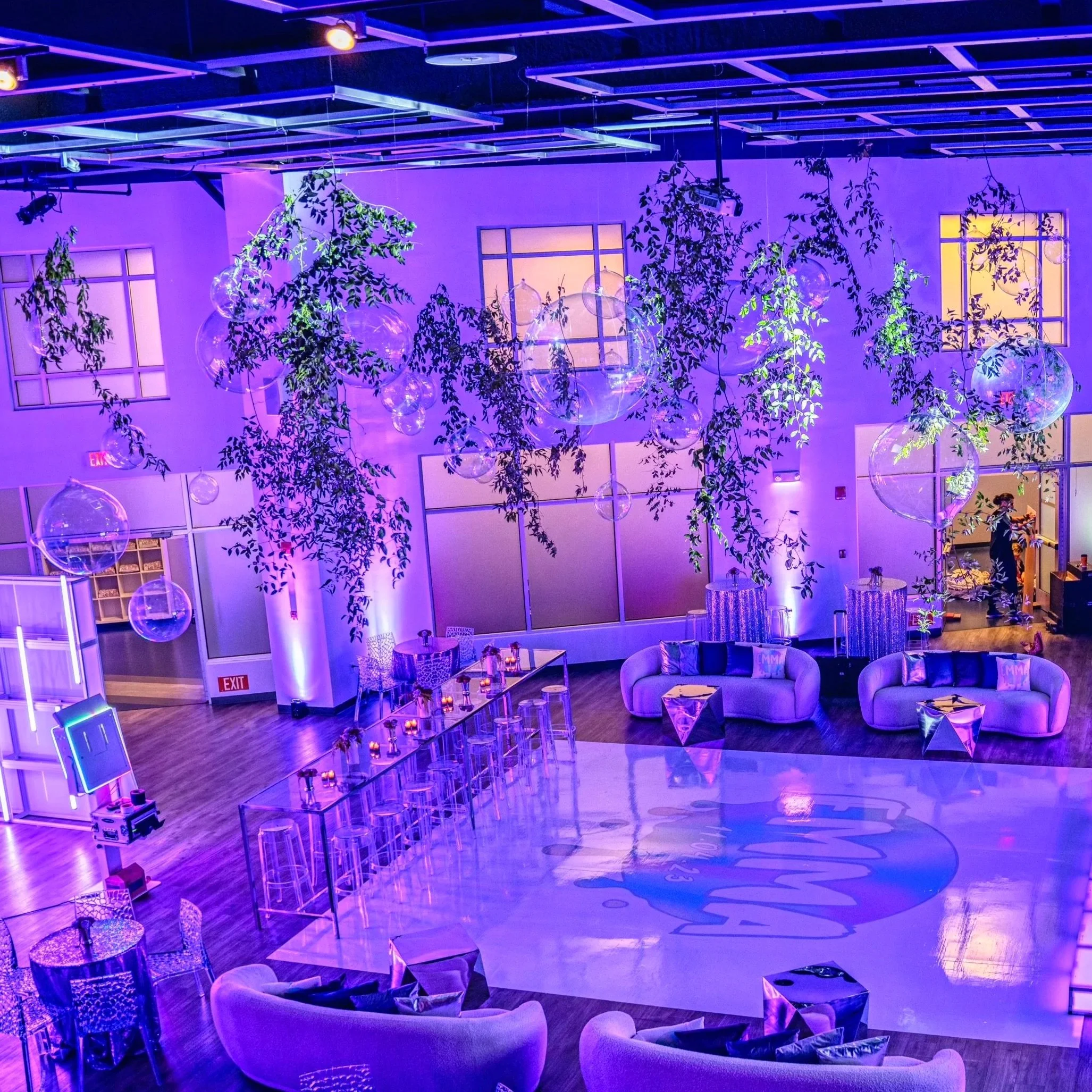A vibrant lounge or event space illuminated with purple and blue lighting. Hanging glass globes and leafy green vines decorate the ceiling. There are modern white couches with throw pillows, a lit-up dance floor with a logo, tables with candles, bar chairs, and a person in the background near the door.