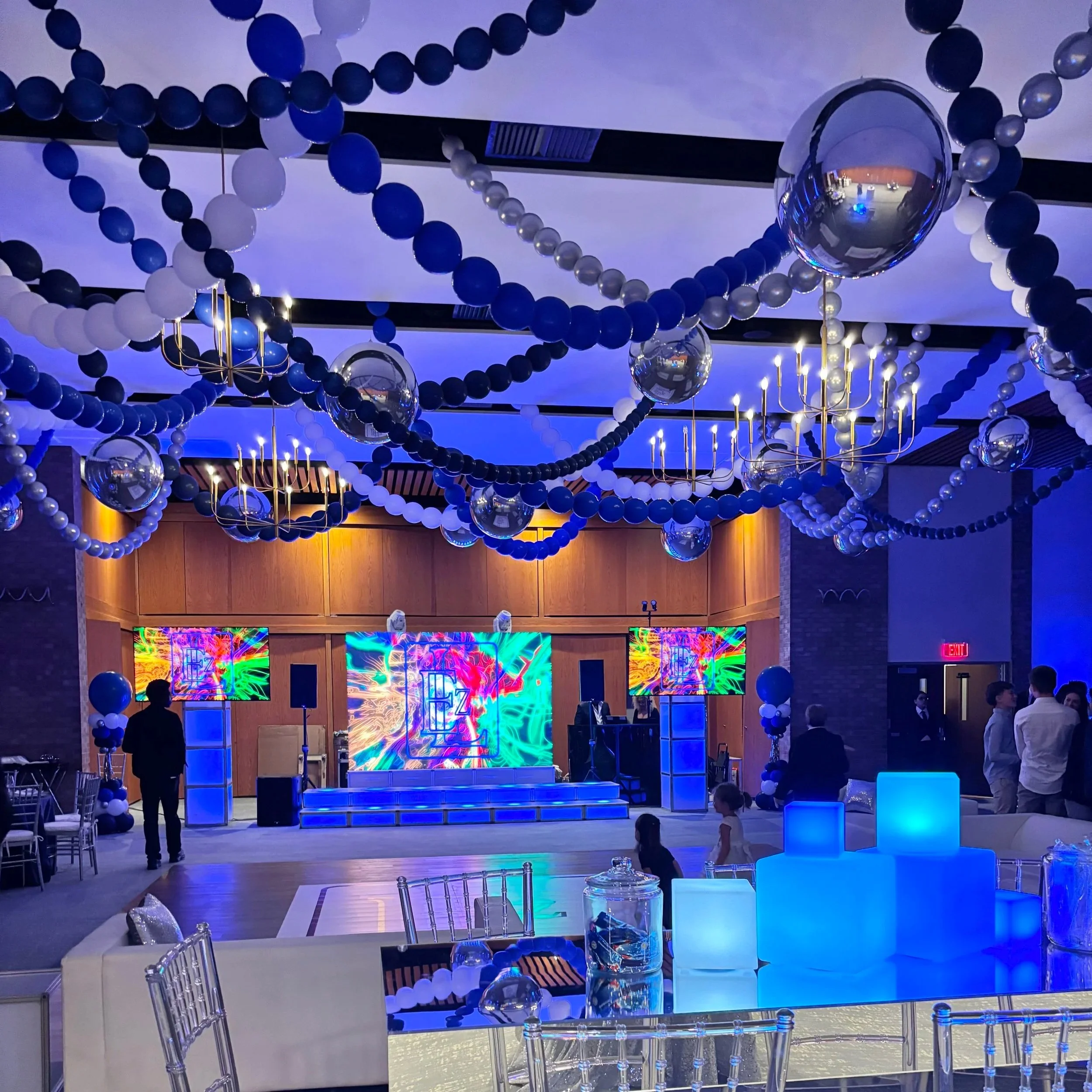 Decorated indoor event space with blue and white balloons, hanging spherical ornaments, and LED displays on stage with people standing and socializing.