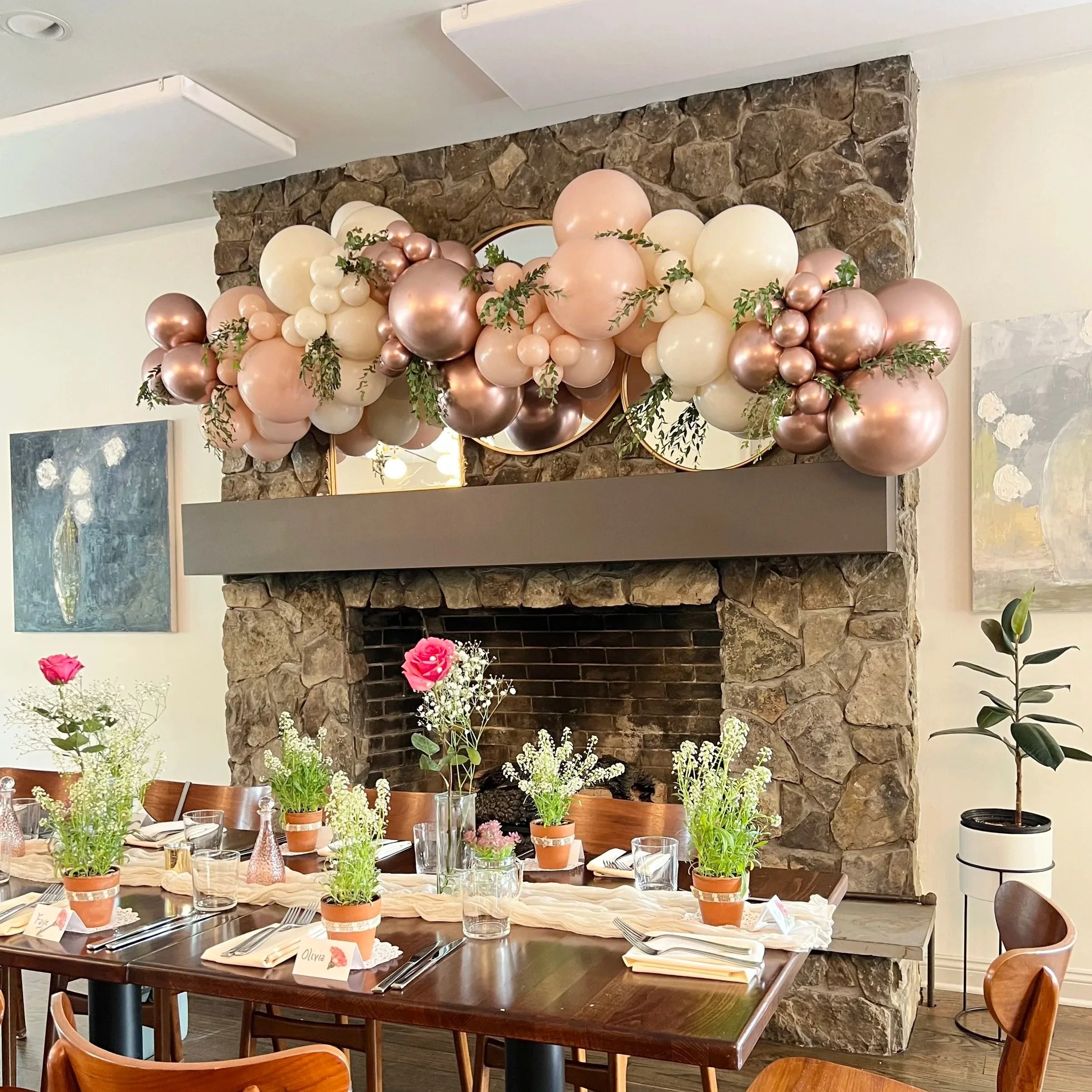 A decorated dining room with a stone fireplace, pink, white, and rose gold balloon garland over the mantel, and a table set with flowers, glassware, and place cards for an event.