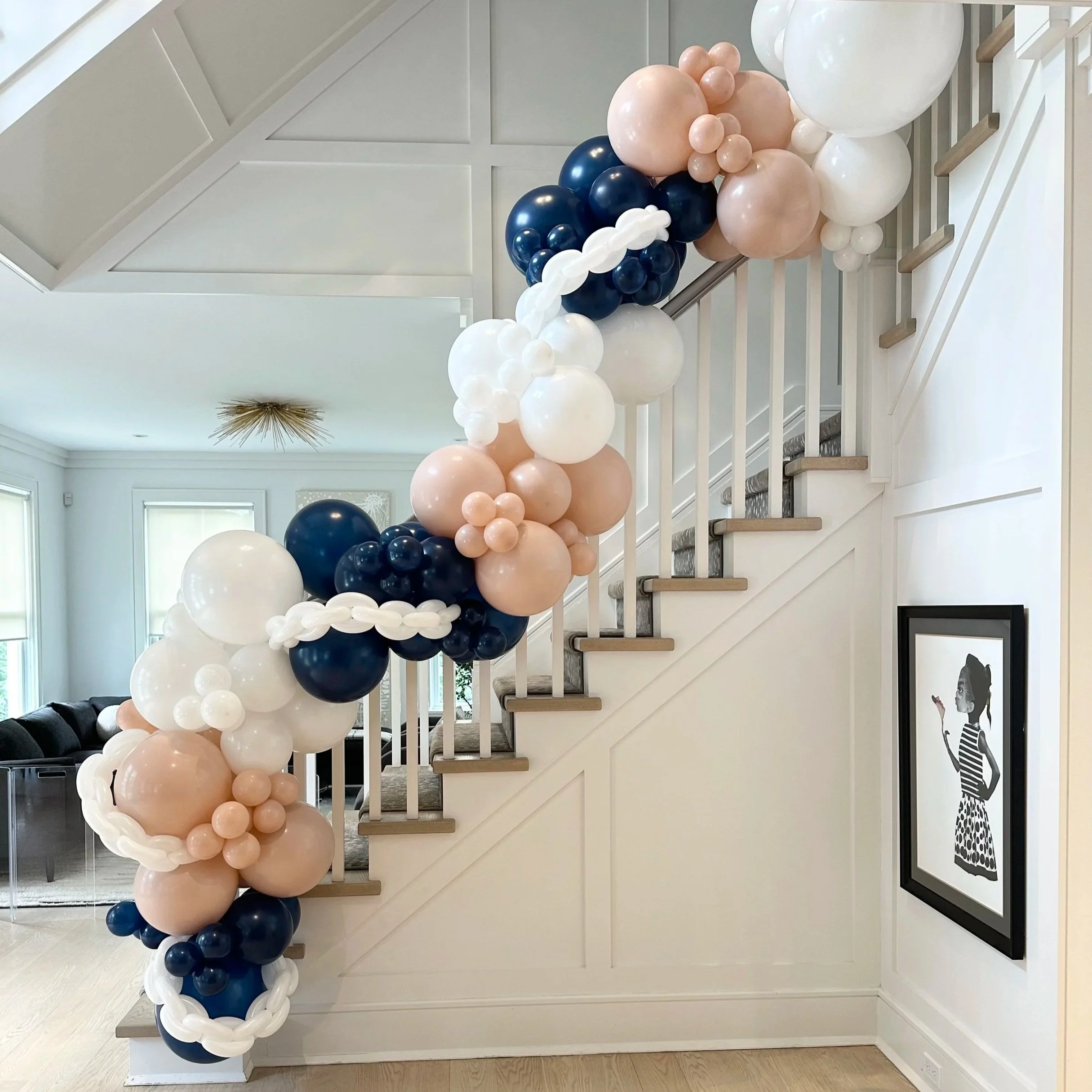 Balloon garland in navy blue, white, blush pink, and pearl beige draped along a staircase railing in a modern living space.