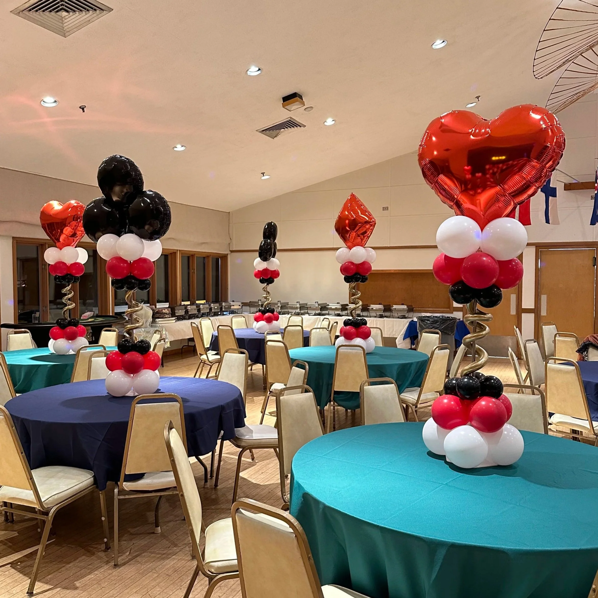 Party room decor with tables covered in teal, navy, and beige tablecloths, each topped with balloon arrangements of hearts in red, white, and black, with large heart-shaped foil balloons and smaller round balloons.