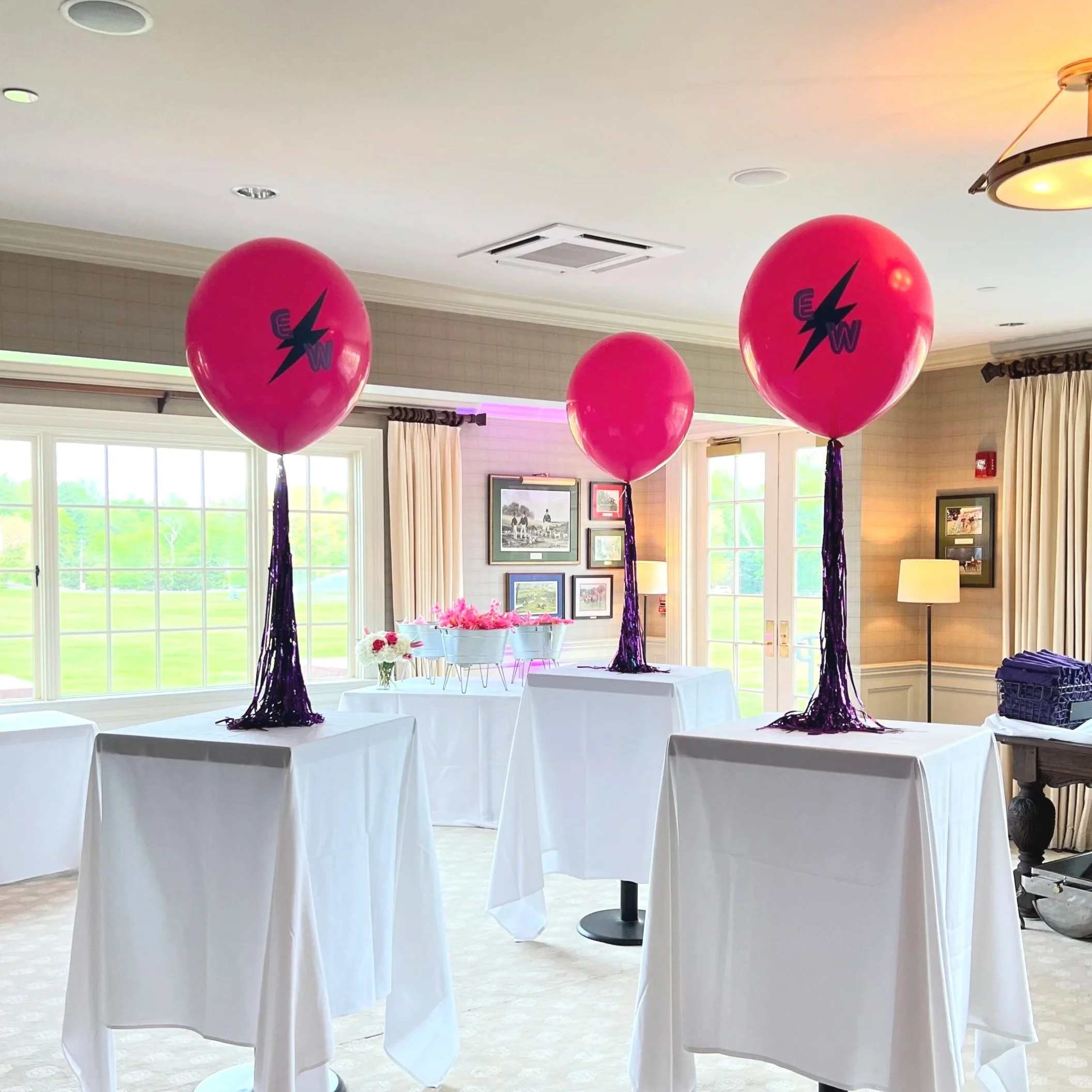 Interior of a decorated event space with three pink balloons on tables, each with purple streamers, and tables with white tablecloths, large windows, and framed pictures on the walls.