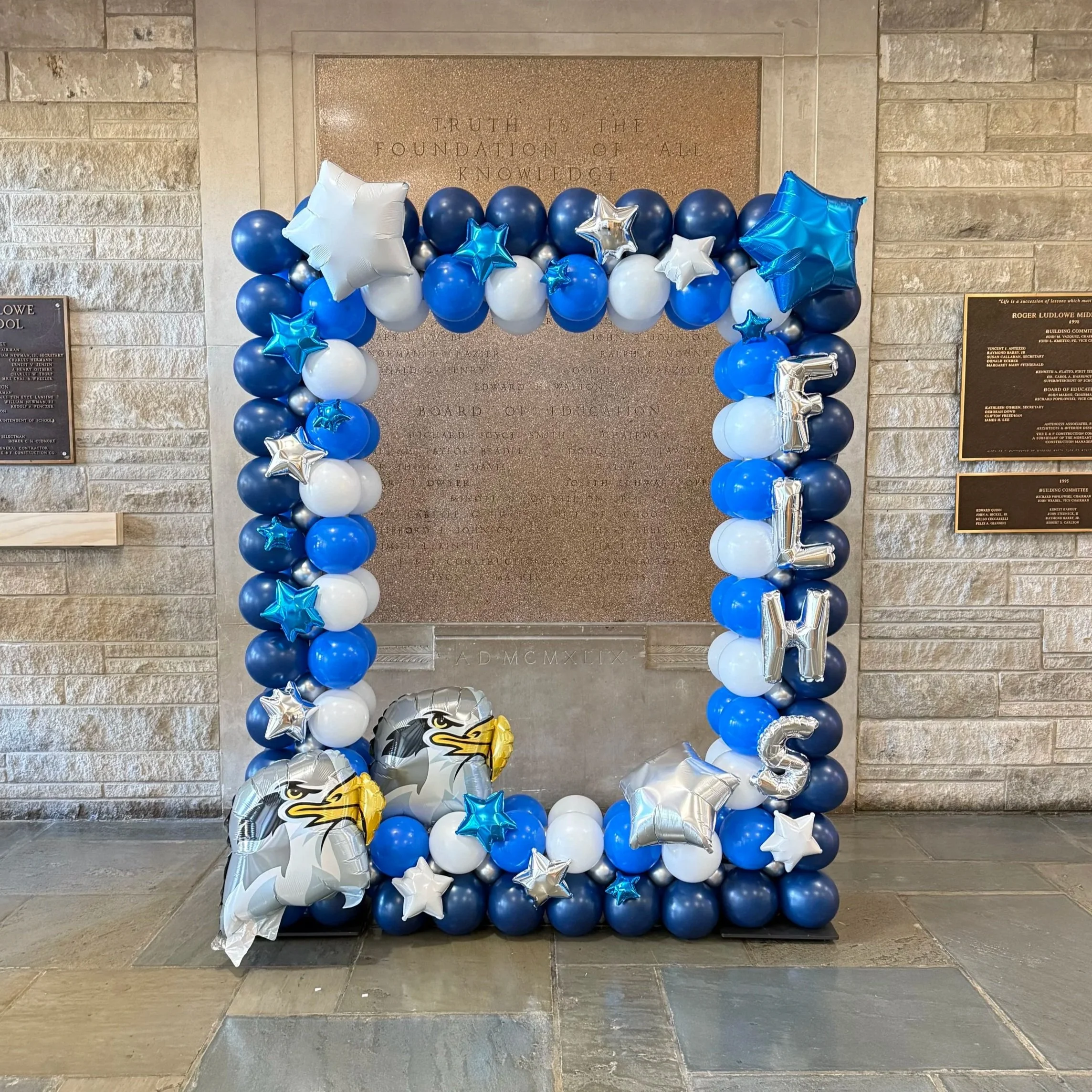 Blue, white, and silver balloons arranged in a decorative arch with eagle and star balloons, with the word "FAMILIES" spelled out vertically on the right side. The background features a stone wall and a bronze plaque.
