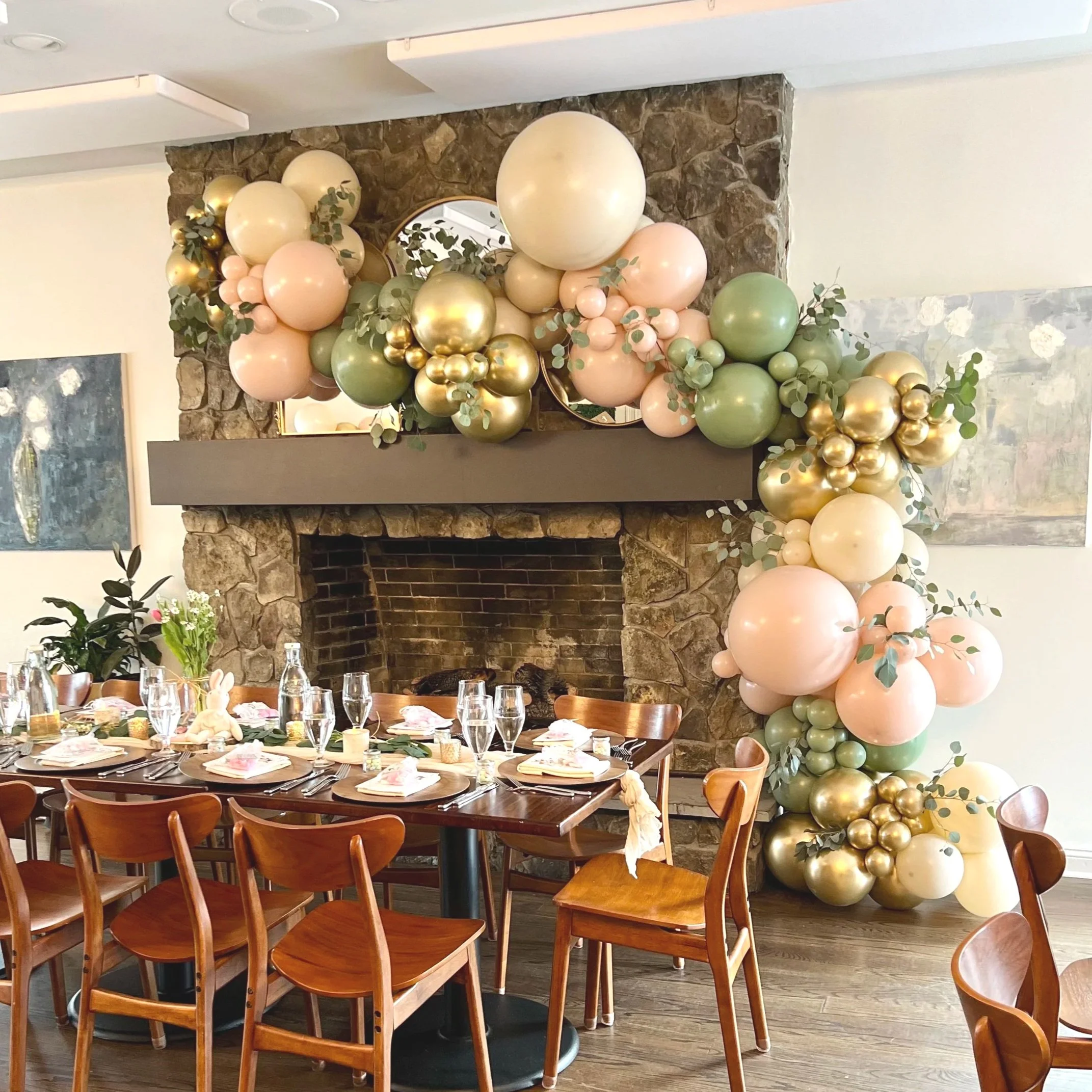Decorative balloon arrangement with pastel pink, green, cream, and gold balloons around a fireplace, with tables set for a celebration or gathering.