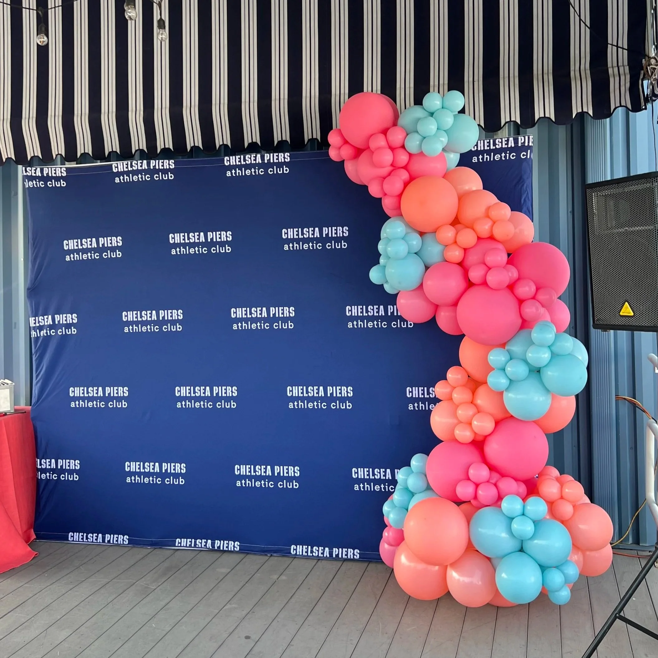 Blue backdrop with white text reading 'Chelsea Piers athletic club' and a large cluster of pastel pink, orange, and blue balloons on the right side.