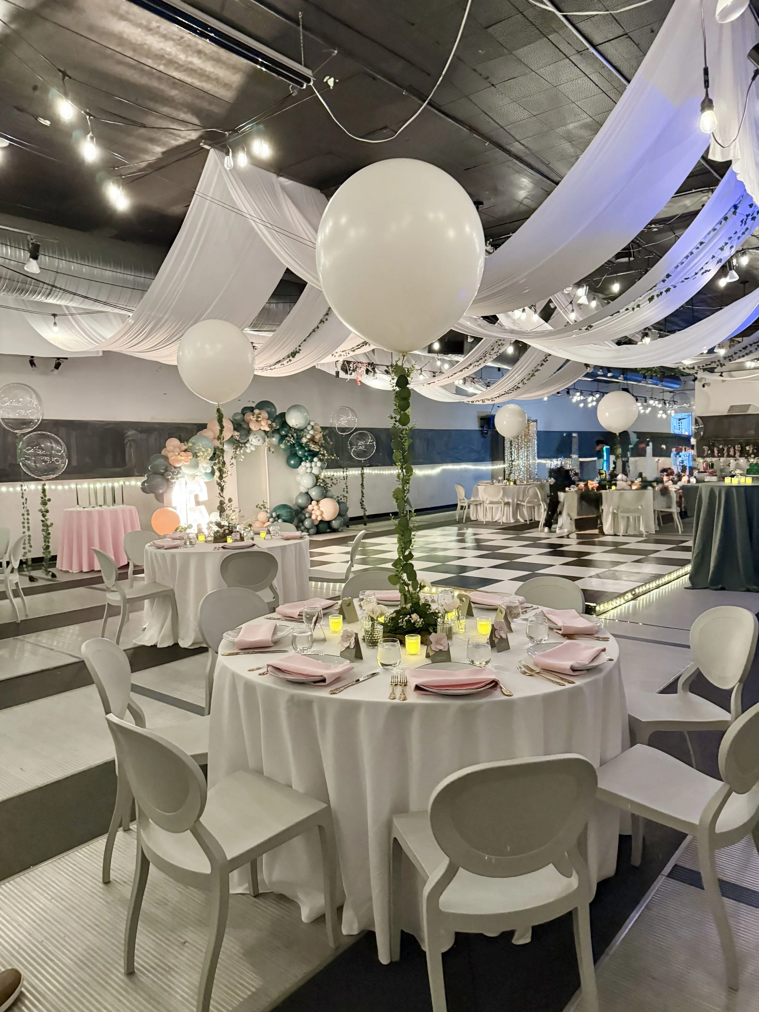 Decorated banquet hall with white draped fabric on the ceiling, large white balloons on long green stems, round tables with white tablecloths, pink napkins, gold cutlery, and small yellow candles, and a balloon arch in the background.