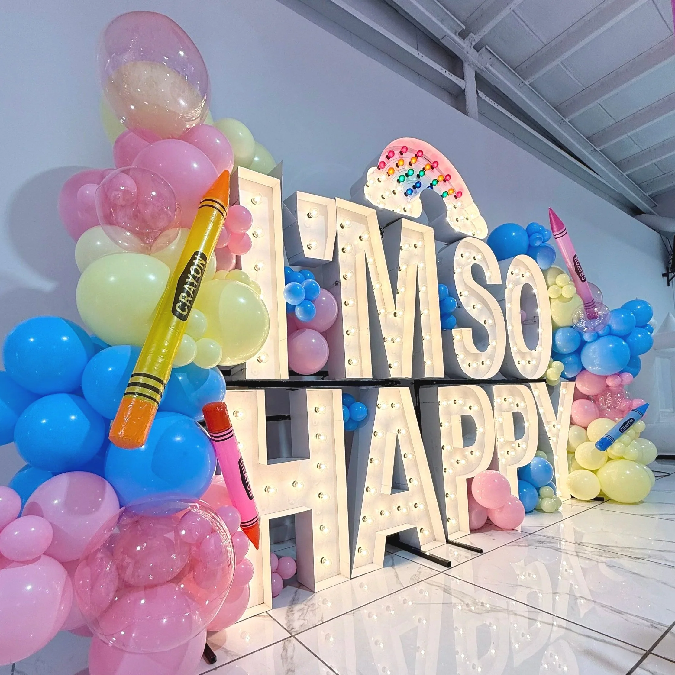 Decorative party sign saying 'It's a MISO Happy HAPP' with large illuminated letters, surrounded by pastel pink, yellow, and blue balloons and large crayon-shaped decorations, with a rainbow-shaped light above.