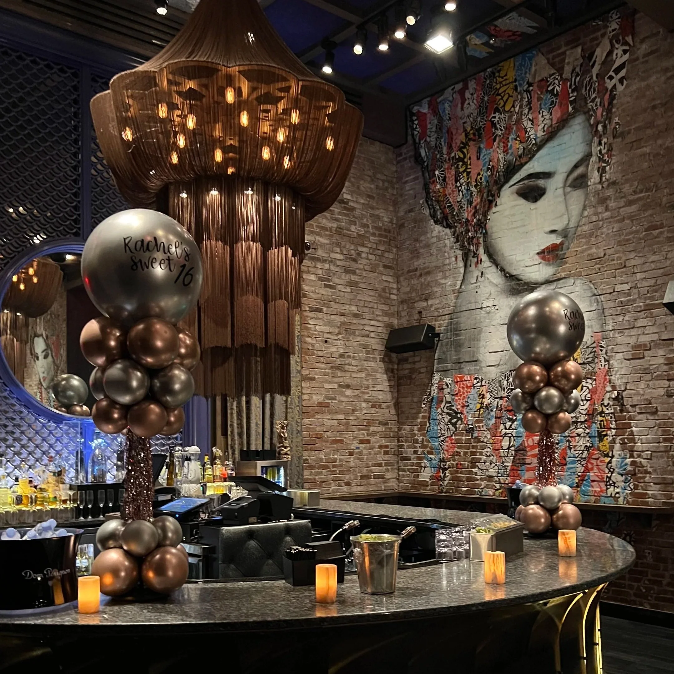 Decorative bar with two balloon arrangements celebrating Rachel's 16th birthday, featuring metallic balloons and a large painting of a woman with colorful headscarf on brick wall.