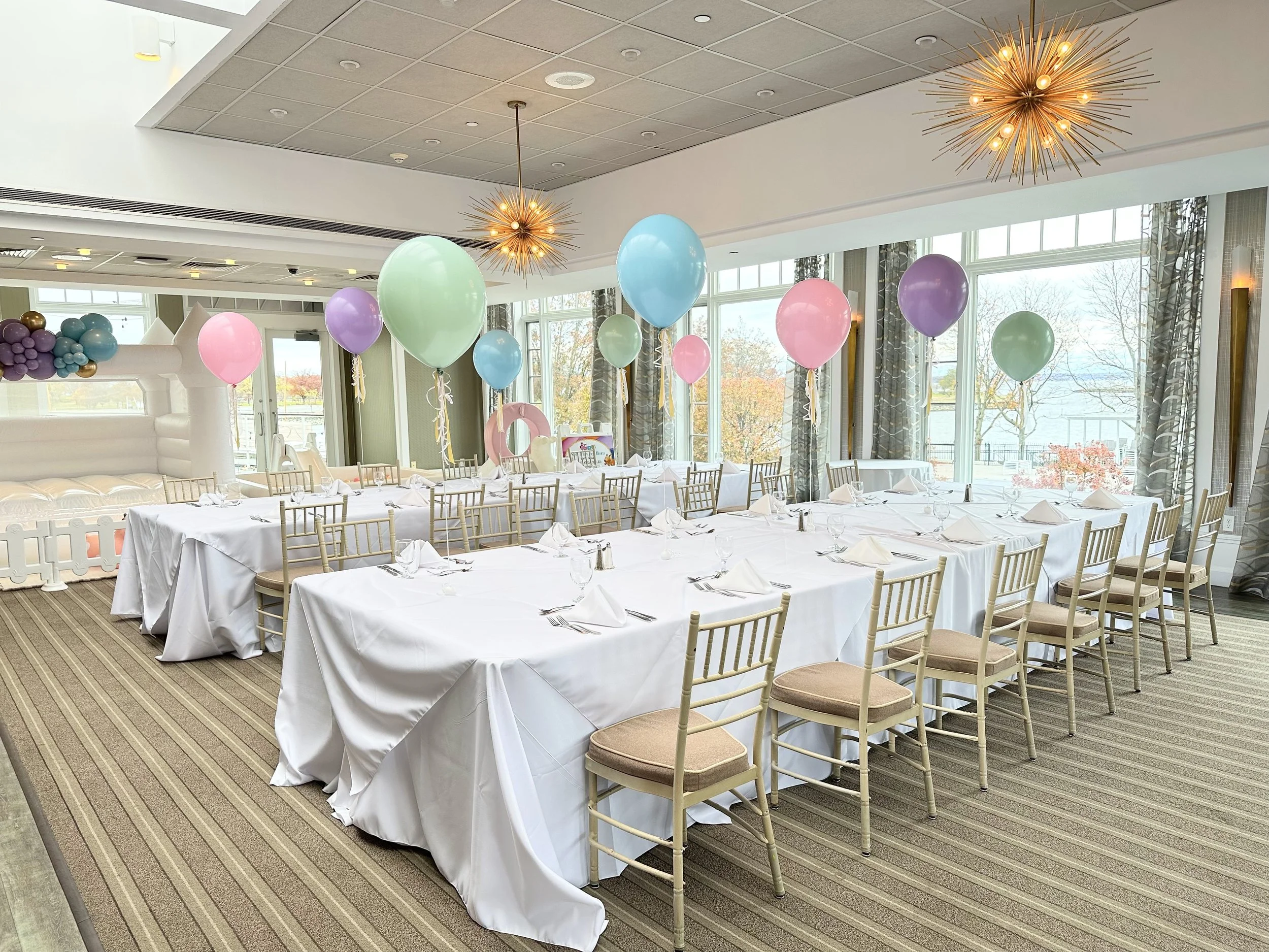 Party setup with long tables covered in white tablecloths, decorated with pastel balloons hanging from the ceiling, and a view of large windows showing an outdoor scene with trees.