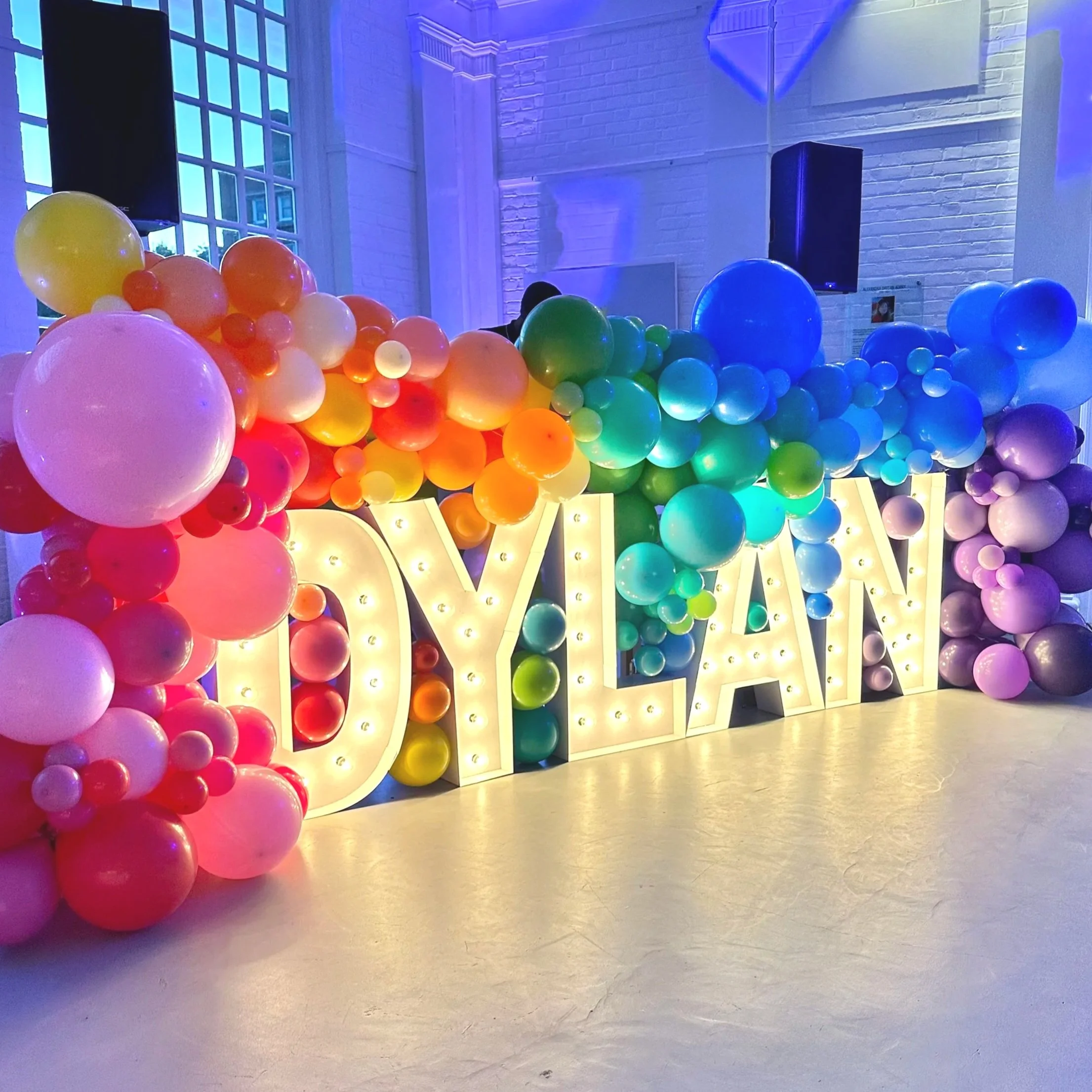 Colorful balloon arch with large lit-up sign spelling 'DYLAYAN' in front, inside a venue with white brick walls and large windows.