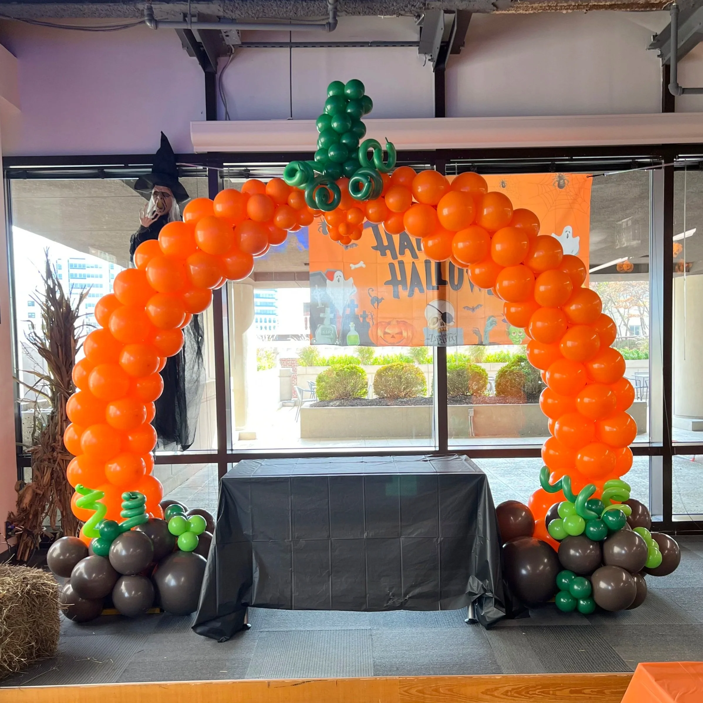 Halloween themed balloon arch with orange, black, green, and gray balloons, and a witch decoration on the left, near a window, with a Halloween banner hanging in the background.