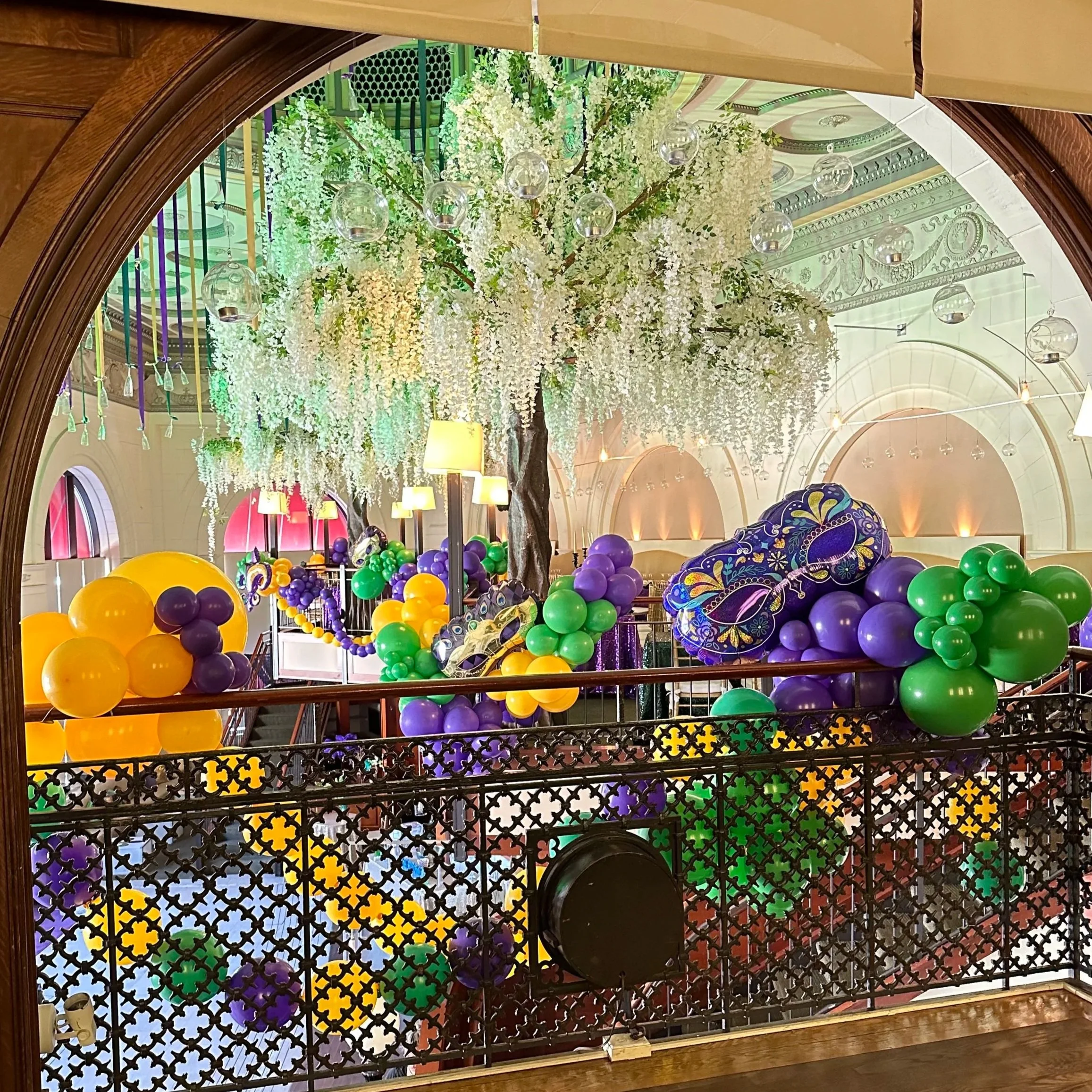 Decorated indoor space with a large white flowering tree, colorful balloons in purple, green, and yellow, decorative masks, and elegant lighting fixtures.