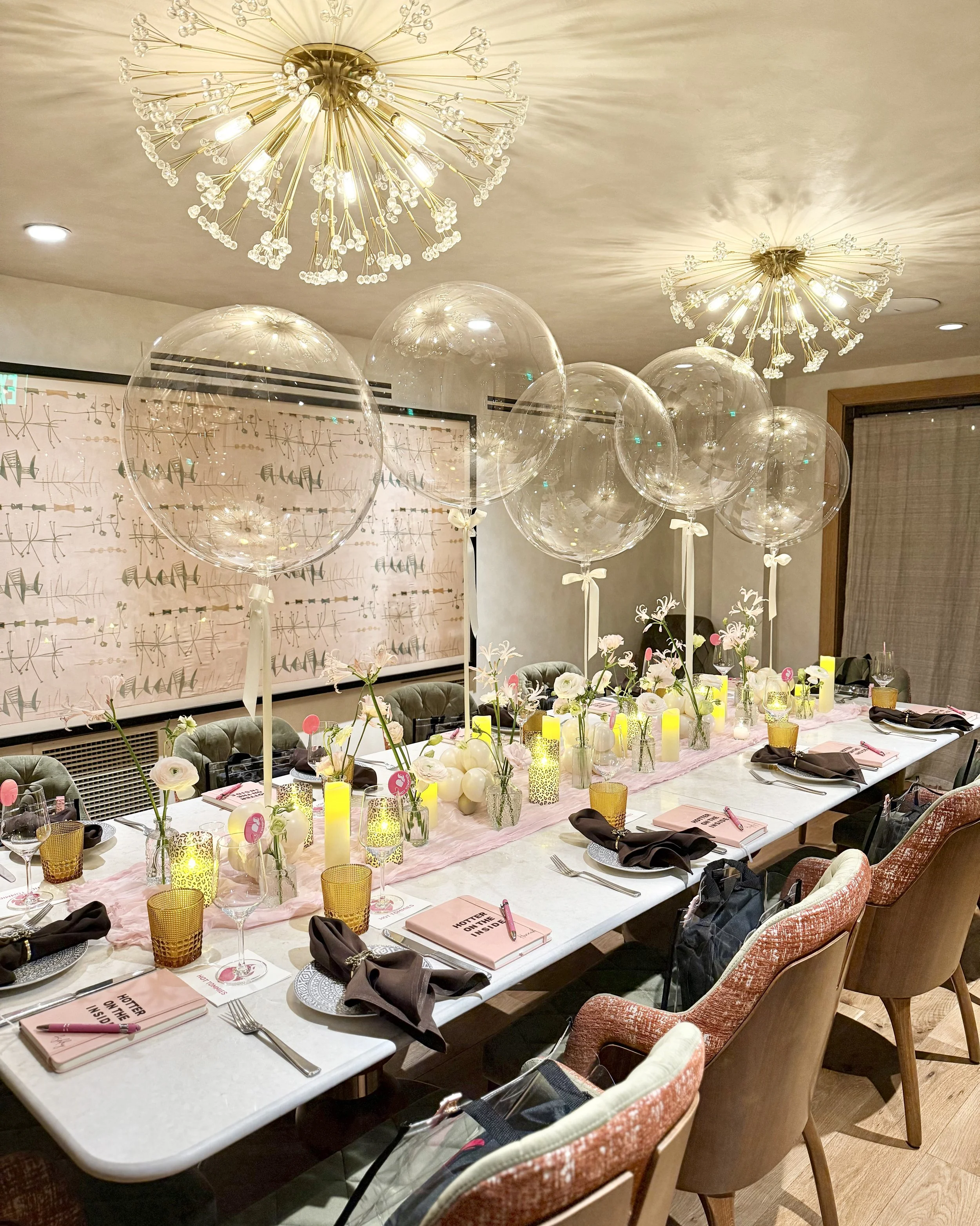A decorated banquet table with balloons, candles, flowers, and place settings, set for a celebration or event in a modern room with ceiling chandeliers.
