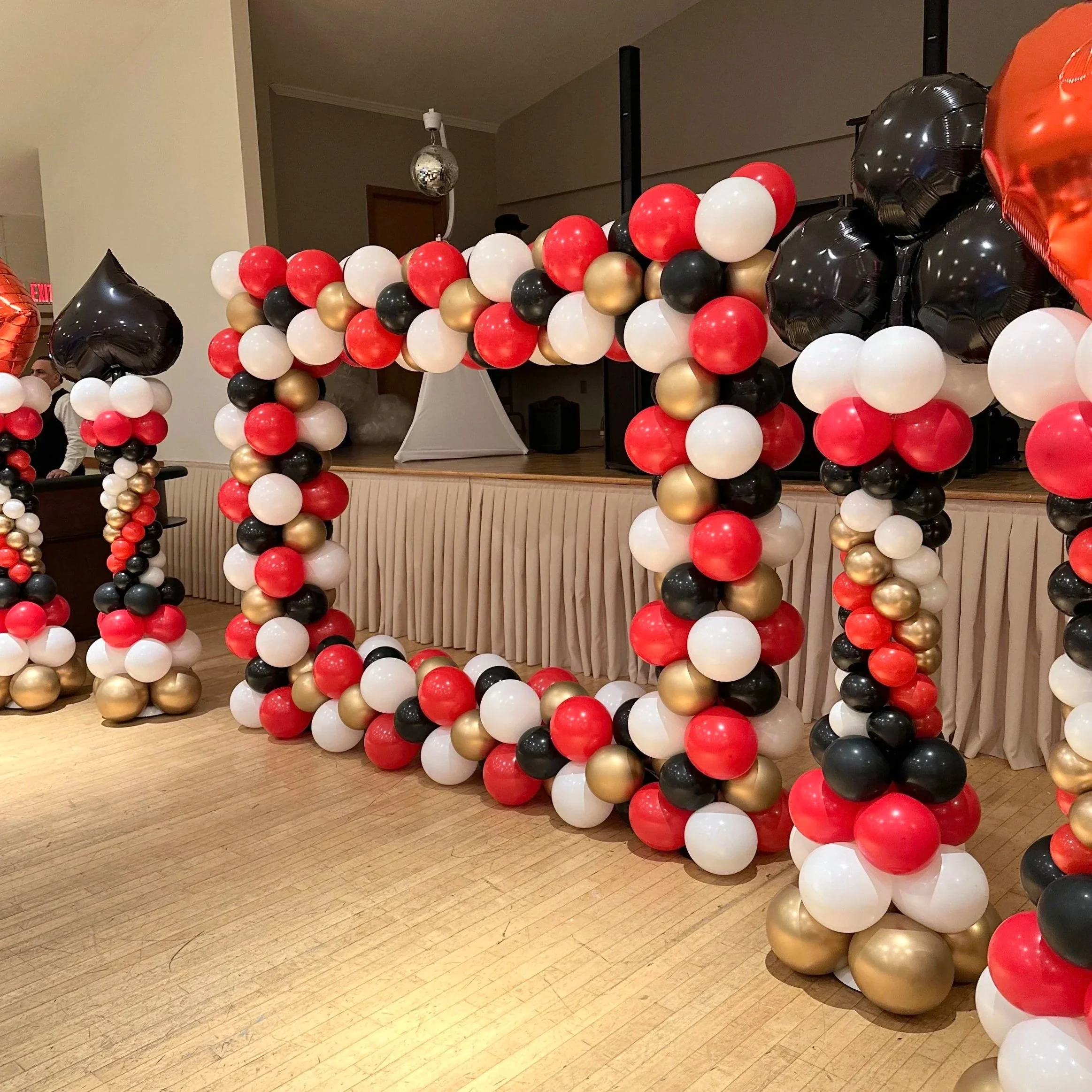 Decorative balloon arch made of red, white, black, and gold balloons, with black heart-shaped balloons on either side, set up at an indoor event.