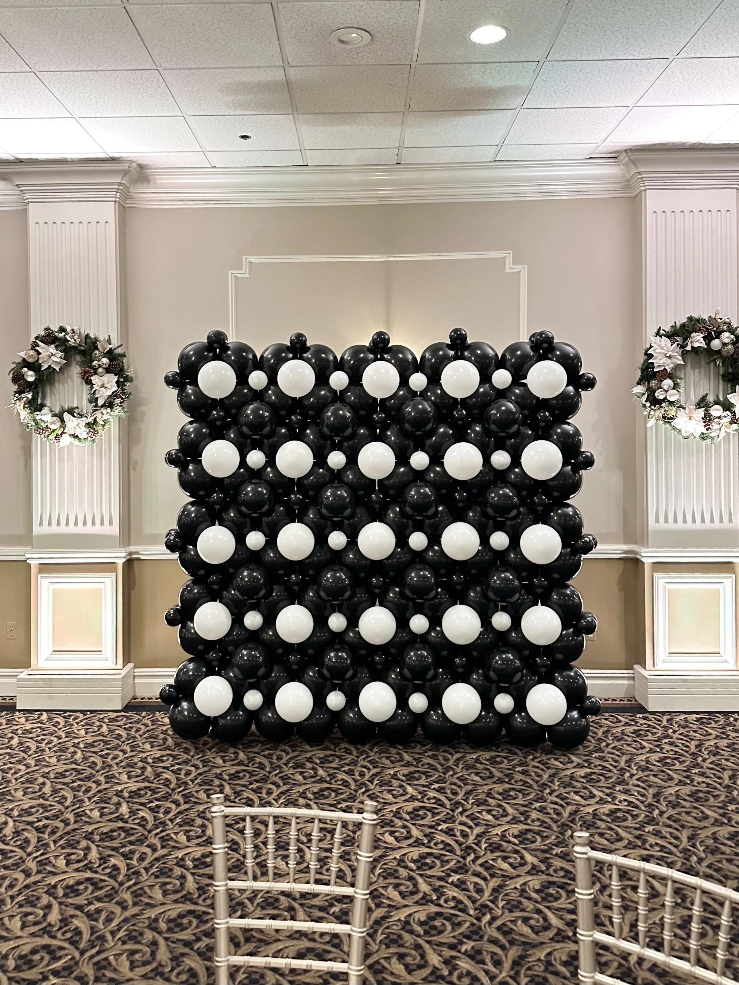 A balloon wall decorated with black and white balloons, flanked by Christmas wreaths on each side, in a decorated event room.