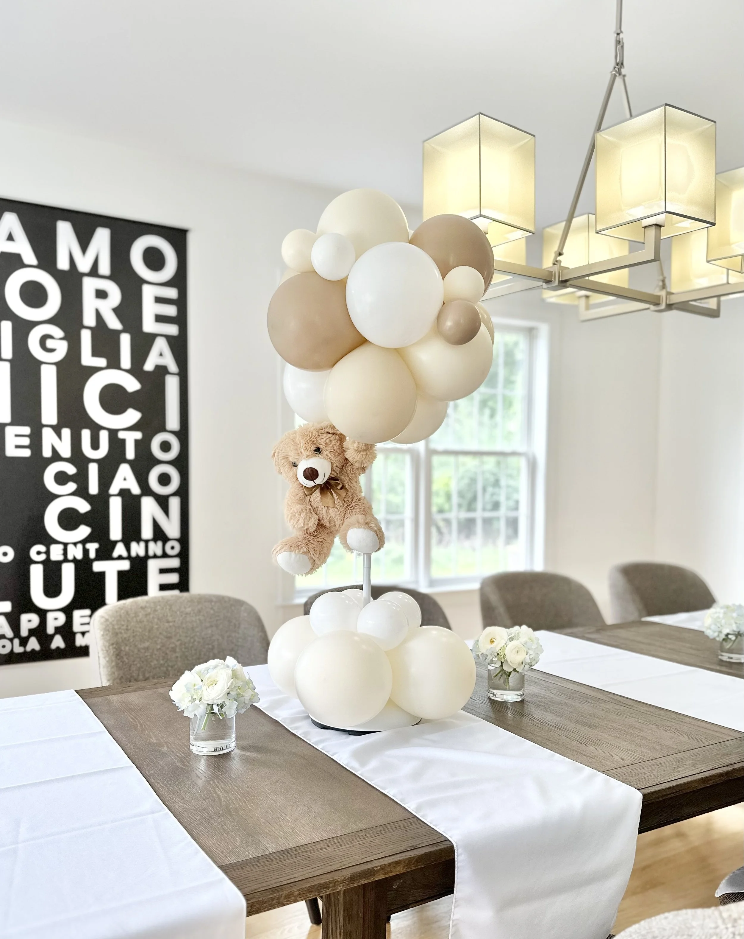 Teddy bear sitting among white, beige, and tan balloons as a table centerpiece in a decorated dining room.