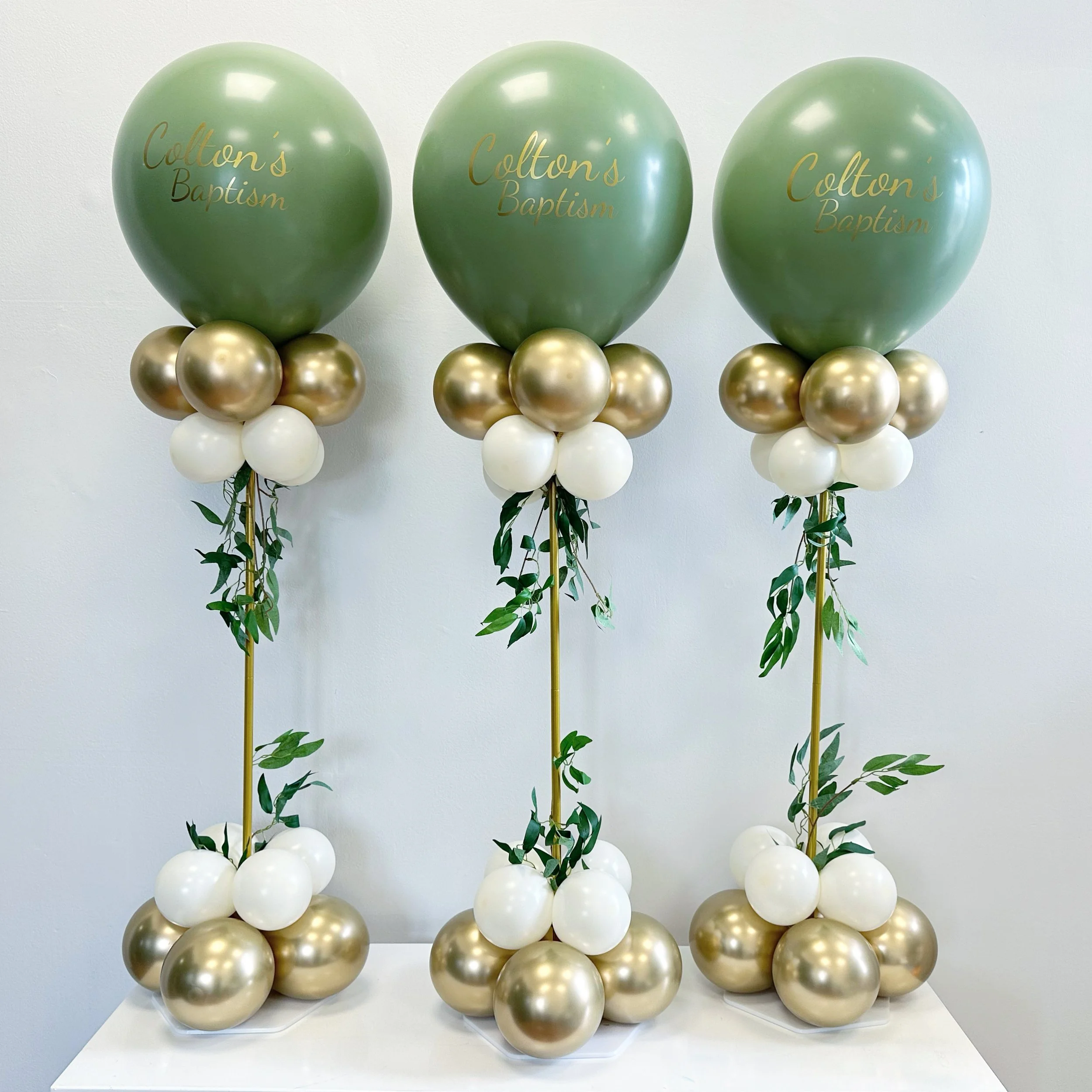 Balloon arrangements for a baptism, featuring large green balloons with 'Colton's Baptism' written on them, surrounded by gold and white balloons, with greenery accents.