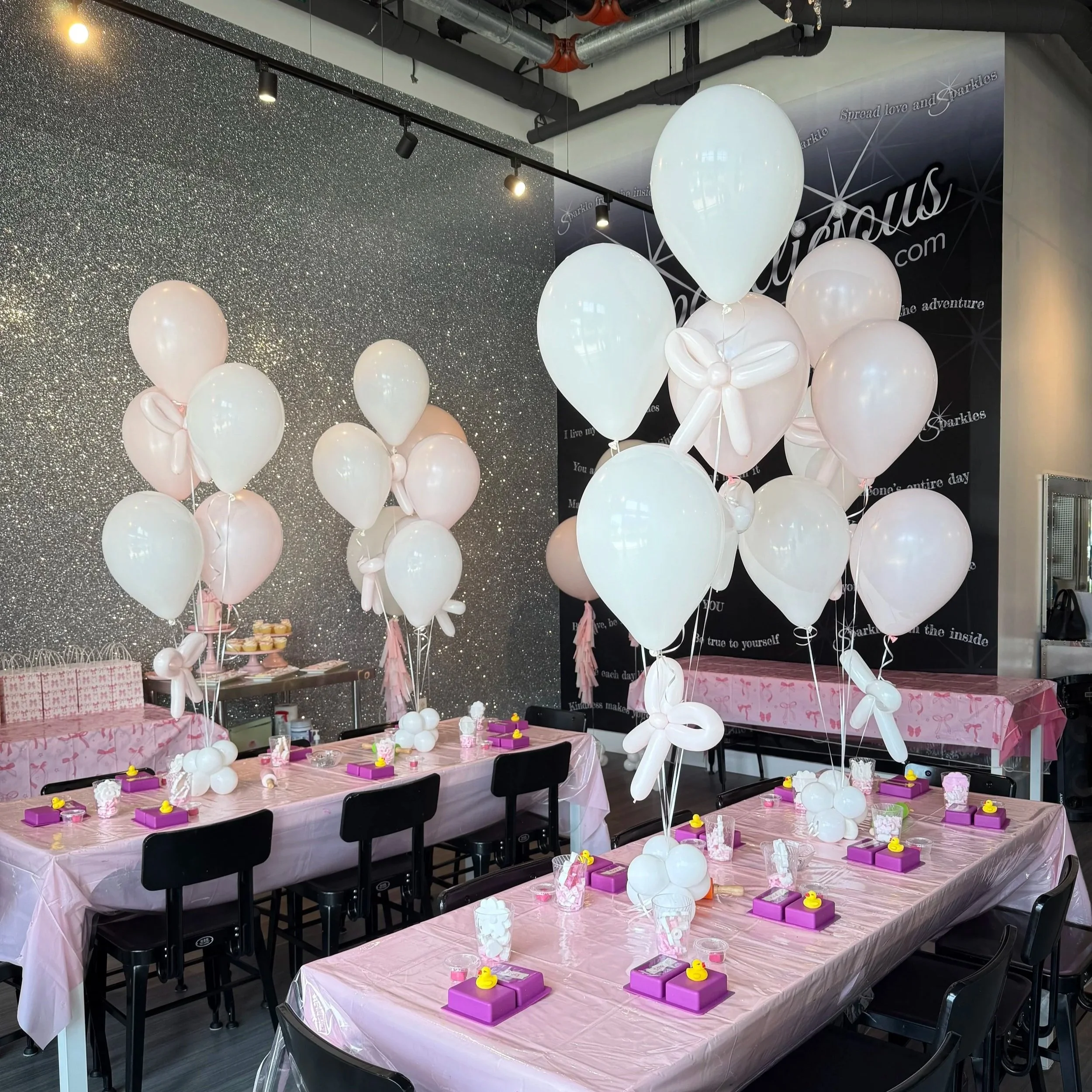 Party room decorated with white and pink balloons, with tables set for a celebration, featuring pink tablecloths, purple boxes, and yellow duck decorations.