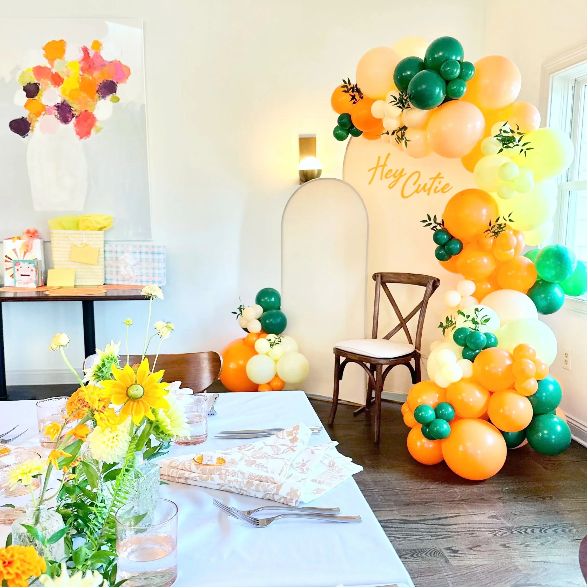 Decorated indoor space with a colorful balloon arch and a table set for a celebration, featuring yellow floral centerpieces, a wall painting of a vase with flowers, and birthday presents.