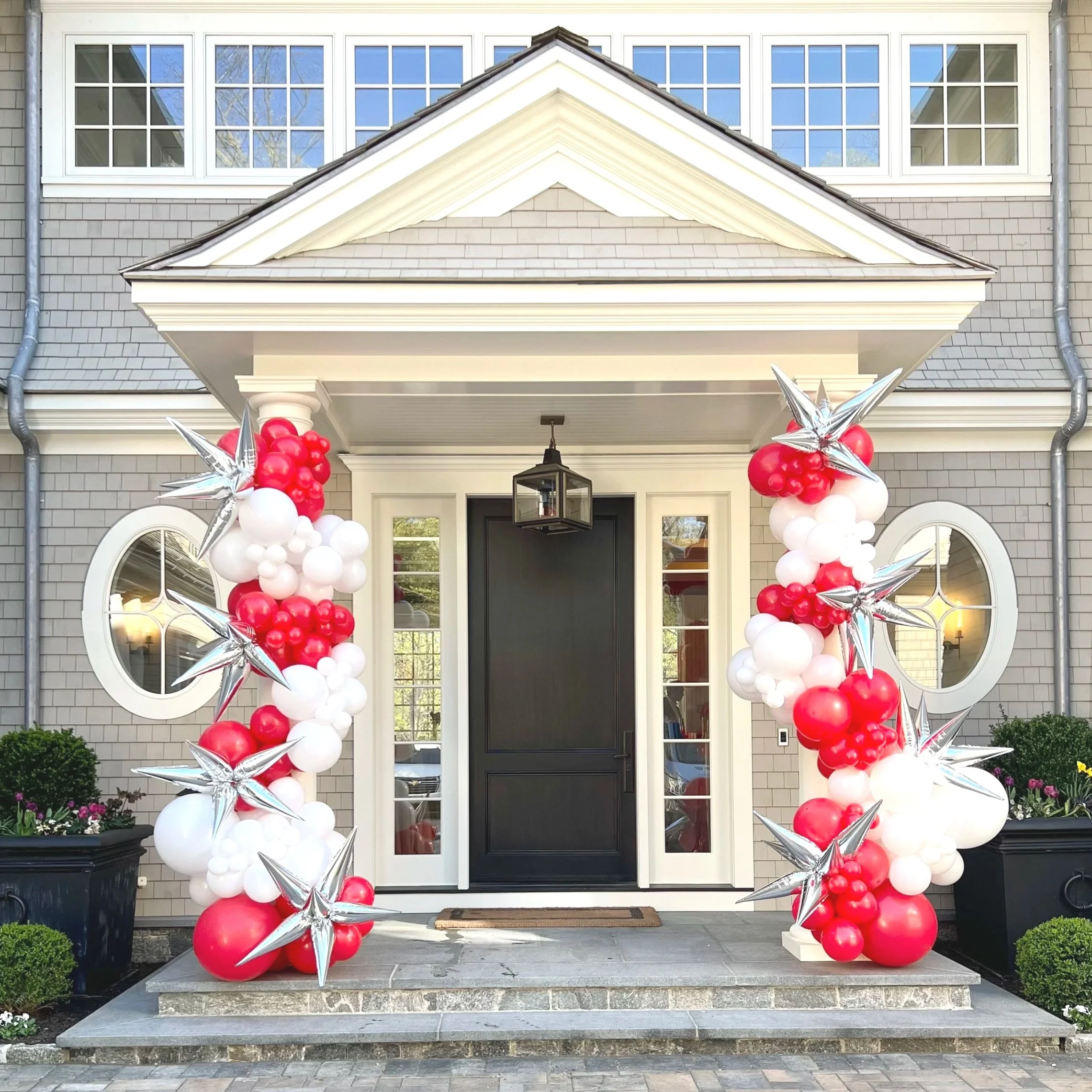 Decorated front porch of a house with red, white, and silver balloons arranged around the door, with silver star-shaped balloons, and a black door with glass panels. House has gray siding, circular windows on sides, and potted plants outside.