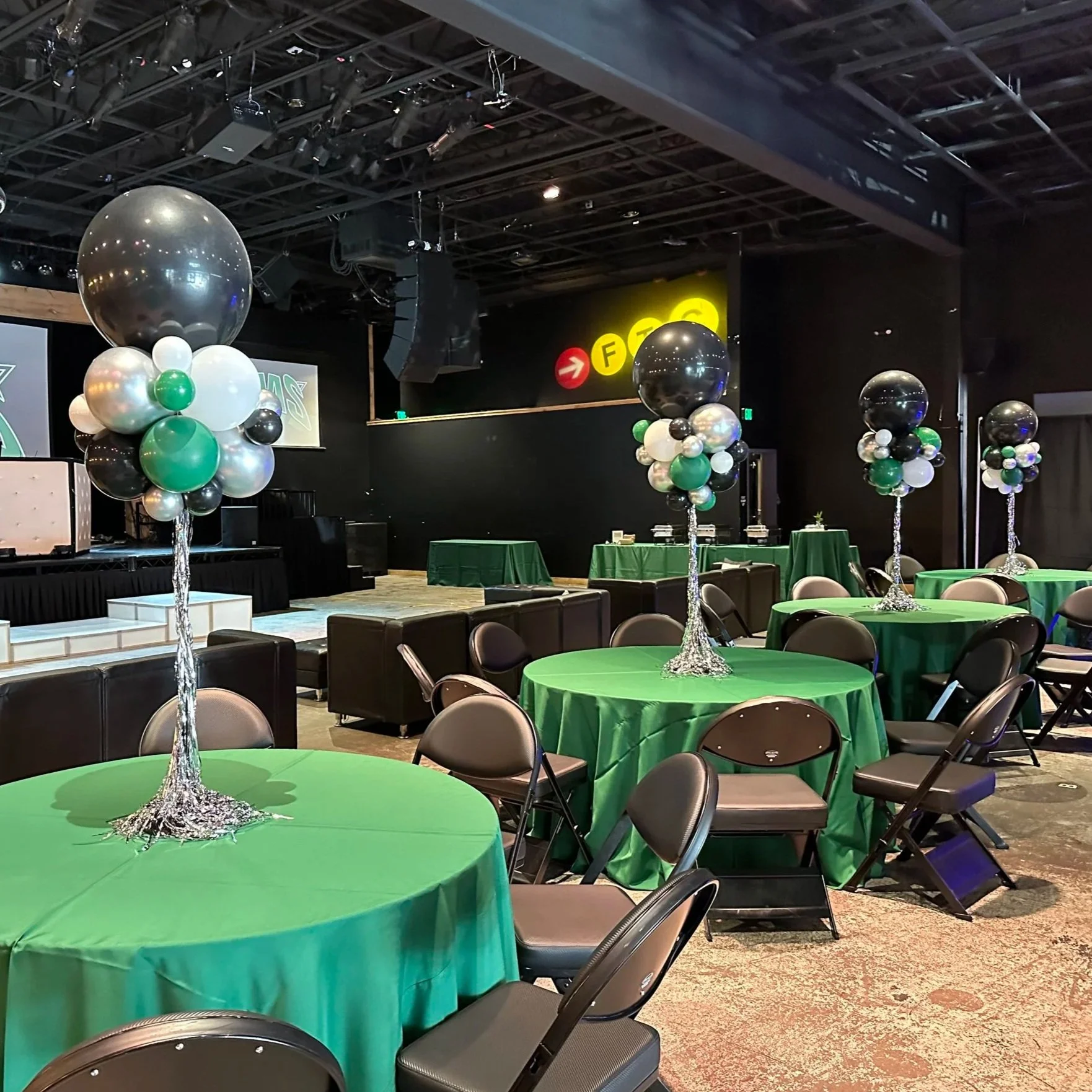 Event space with round tables covered in green tablecloths, each decorated with large clusters of black, white, silver, and green balloons on silver stands, and a stage with a black backdrop and a sound system.