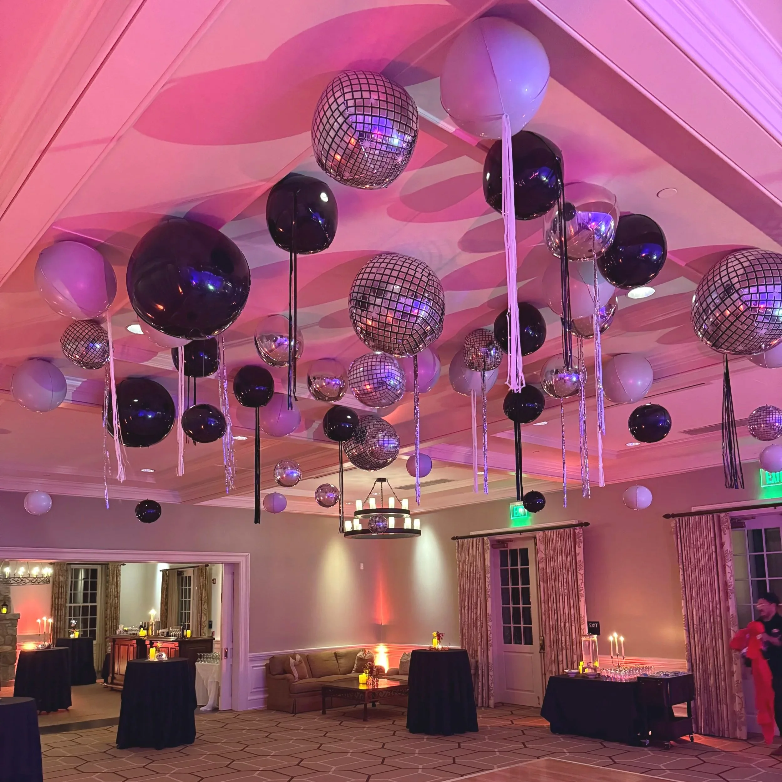Party room decorated with black, white, and mirrored disco ball balloons hanging from the ceiling, some with long hanging ribbons, with dim lighting and candles on tables.