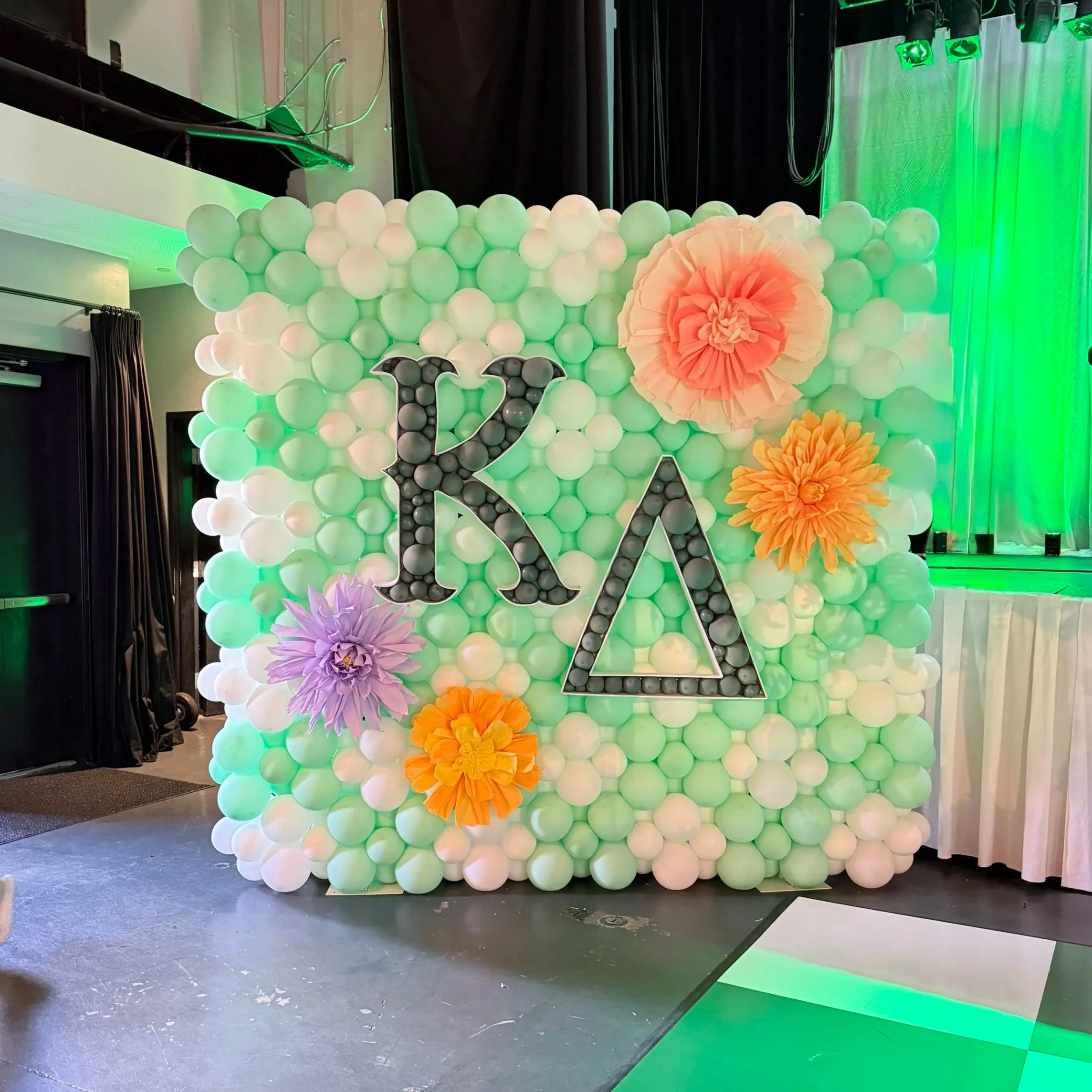 Decorative balloon wall with white and pastel-colored balloons, large paper flowers, and the name "Kara" displayed with light-up letters.