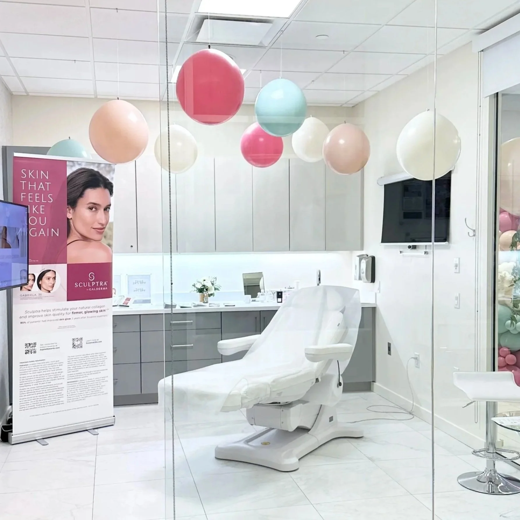 A modern medical or aesthetic office room with a white adjustable treatment chair, colorful balloons hanging from the ceiling, and a large poster featuring a woman's face on the left side.
