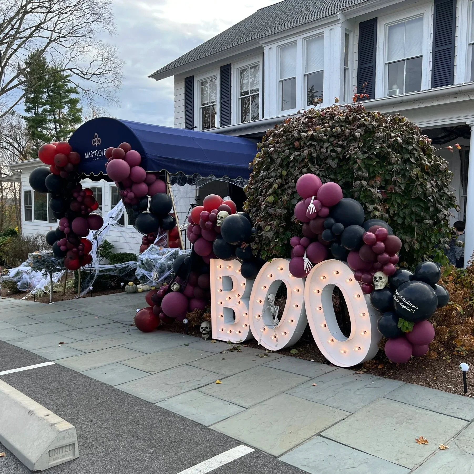 Halloween-themed porch decoration with a large sign that says 'BOO' illuminated with lightbulbs, surrounded by purple, black, and burgundy balloons, some skull decorations, and white spider web decorations on the house and bushes.