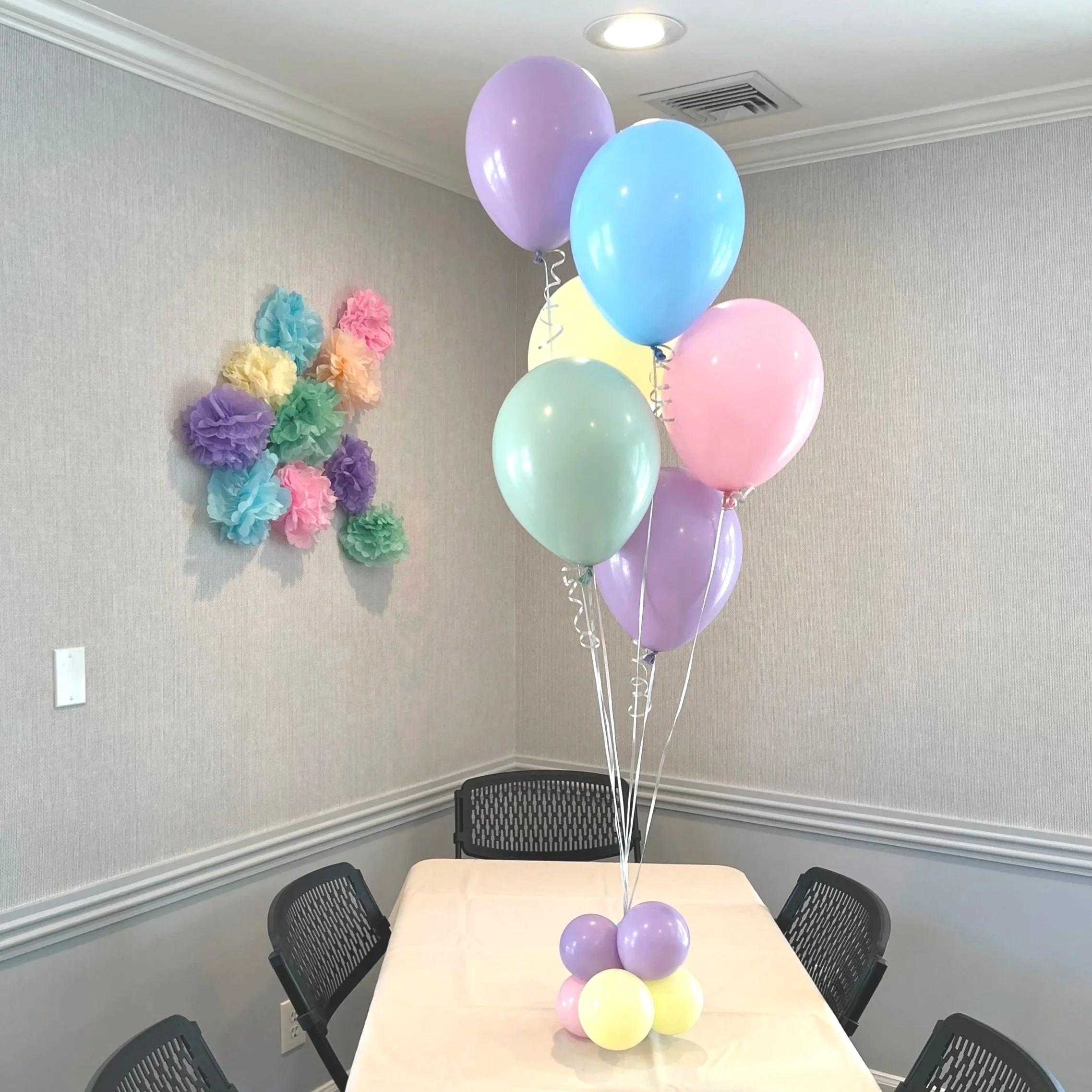 Colorful pastel balloons tied together on a table in a decorated room with tissue paper pom-poms on the wall.