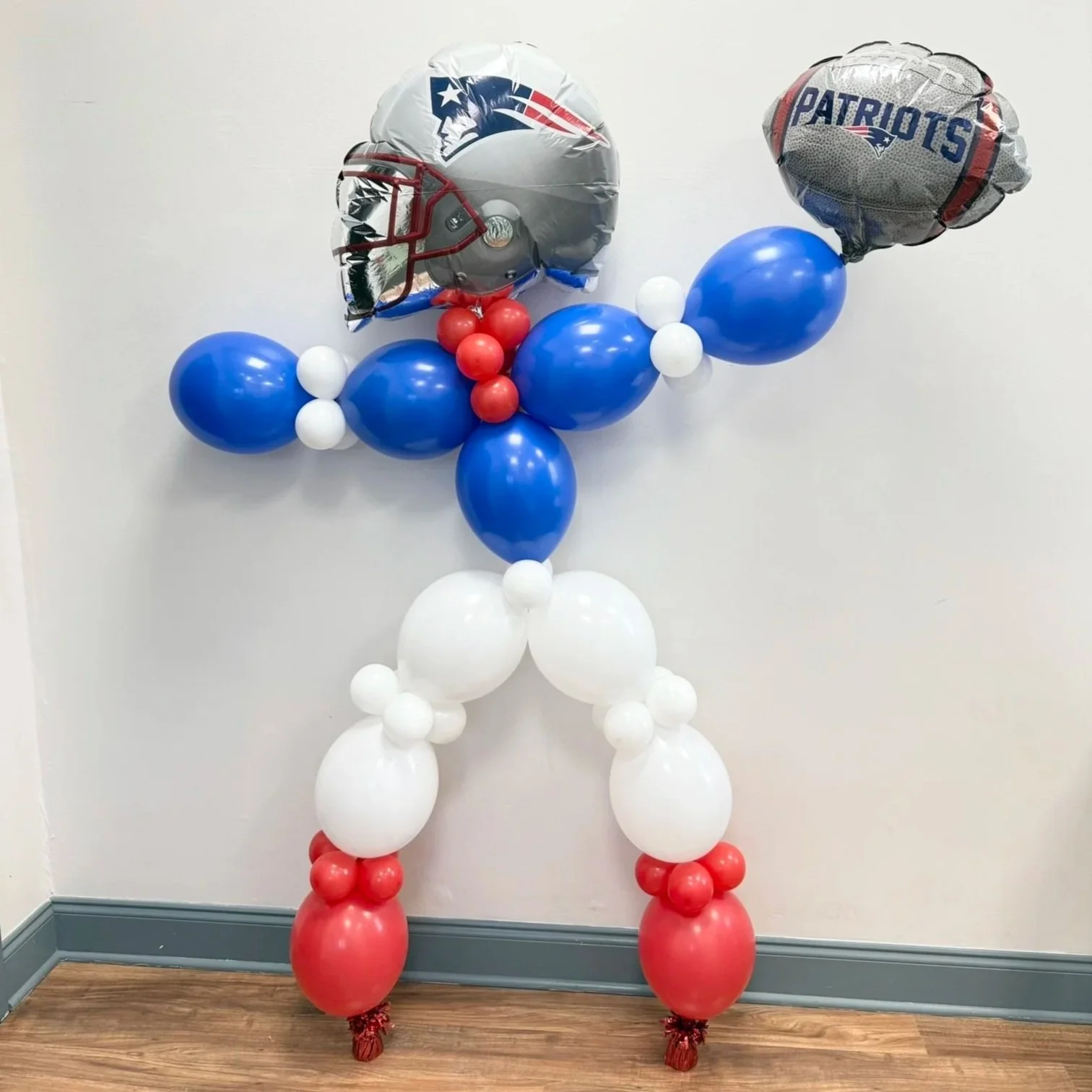 Balloon sculpture of a football player in Patriots team colors, with a Patriots themed helmet and football, against a plain wall.