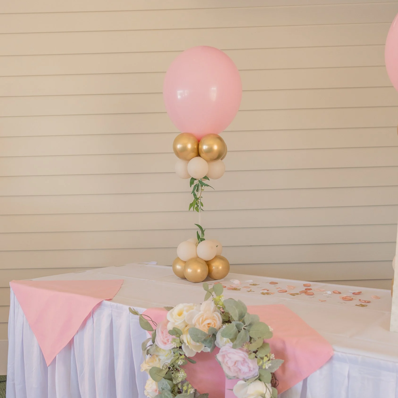 A decorated table with pink and white cloth and a bouquet of flowers, a balloon centerpiece with pink, white, and gold balloons, green leaves, and scattered pink and gold confetti.
