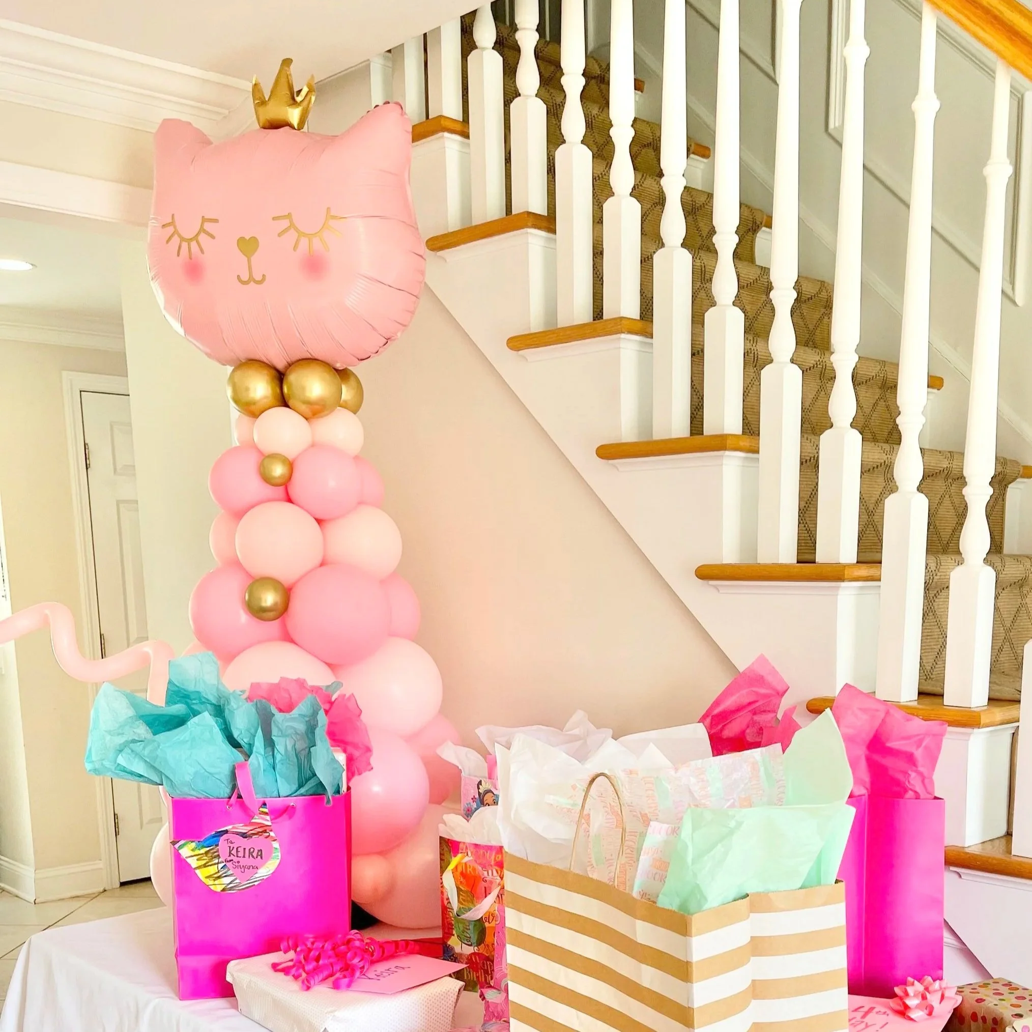 Pink balloon arrangement with a cat face shape and gold crown, pink, white, and gold balloons, gift bags and wrapped presents at a birthday celebration under a staircase.
