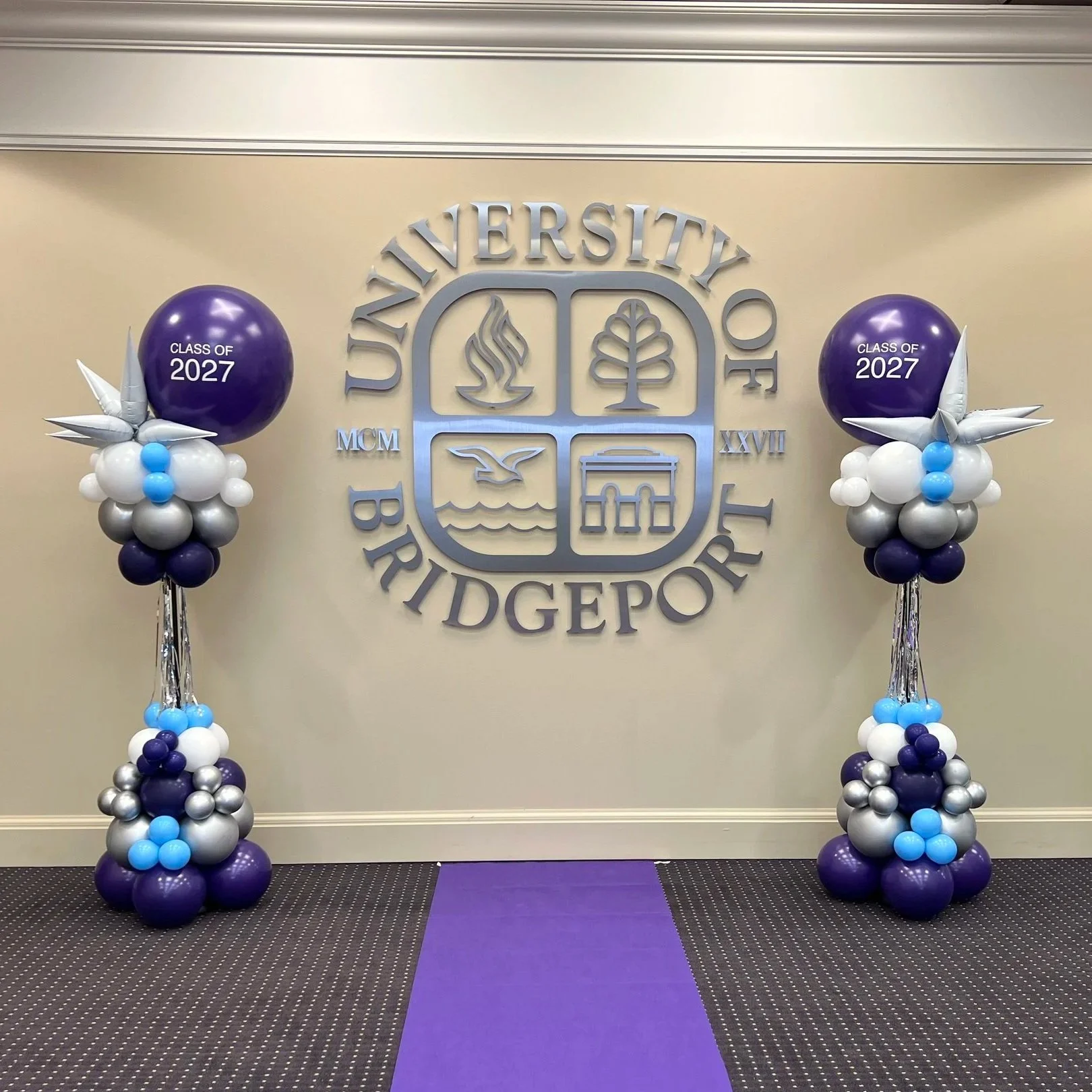 Decorative balloons on either side of a university emblem, with a purple carpet in the center. The balloons are purple, white, black, silver, and blue, with large purple balloons reading 'Class of 2027' on top, and silver, white, black, and blue smaller balloons underneath.