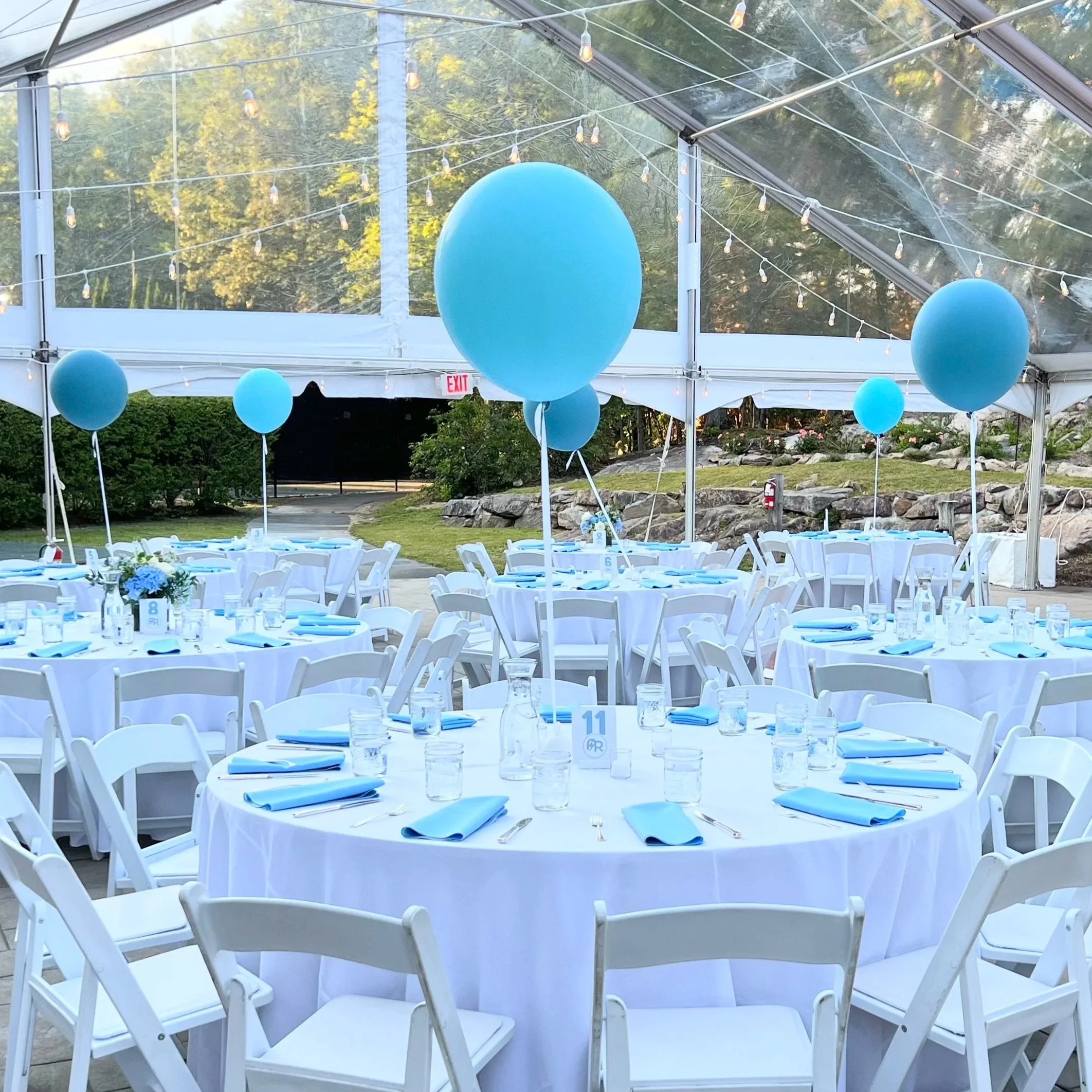 Outdoor event tent with white round tables decorated with blue napkins, glasses, and centerpieces, blue balloons attached to the tables, and string lights overhead, set in a garden area.