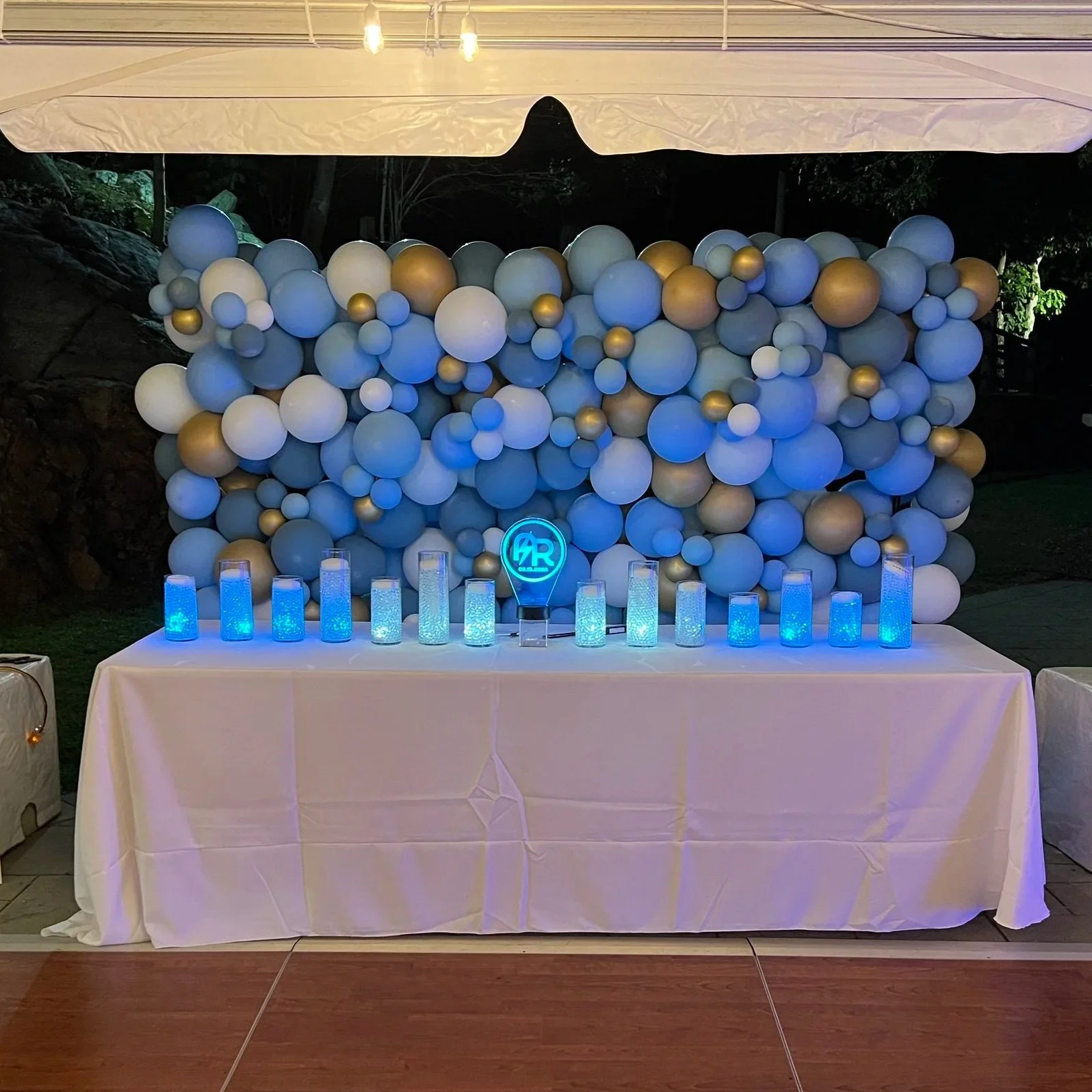 A table with blue illuminated candles and a neon sign reading 'AR' in front of a backdrop of blue, white, and gold balloons at an outdoor night event.