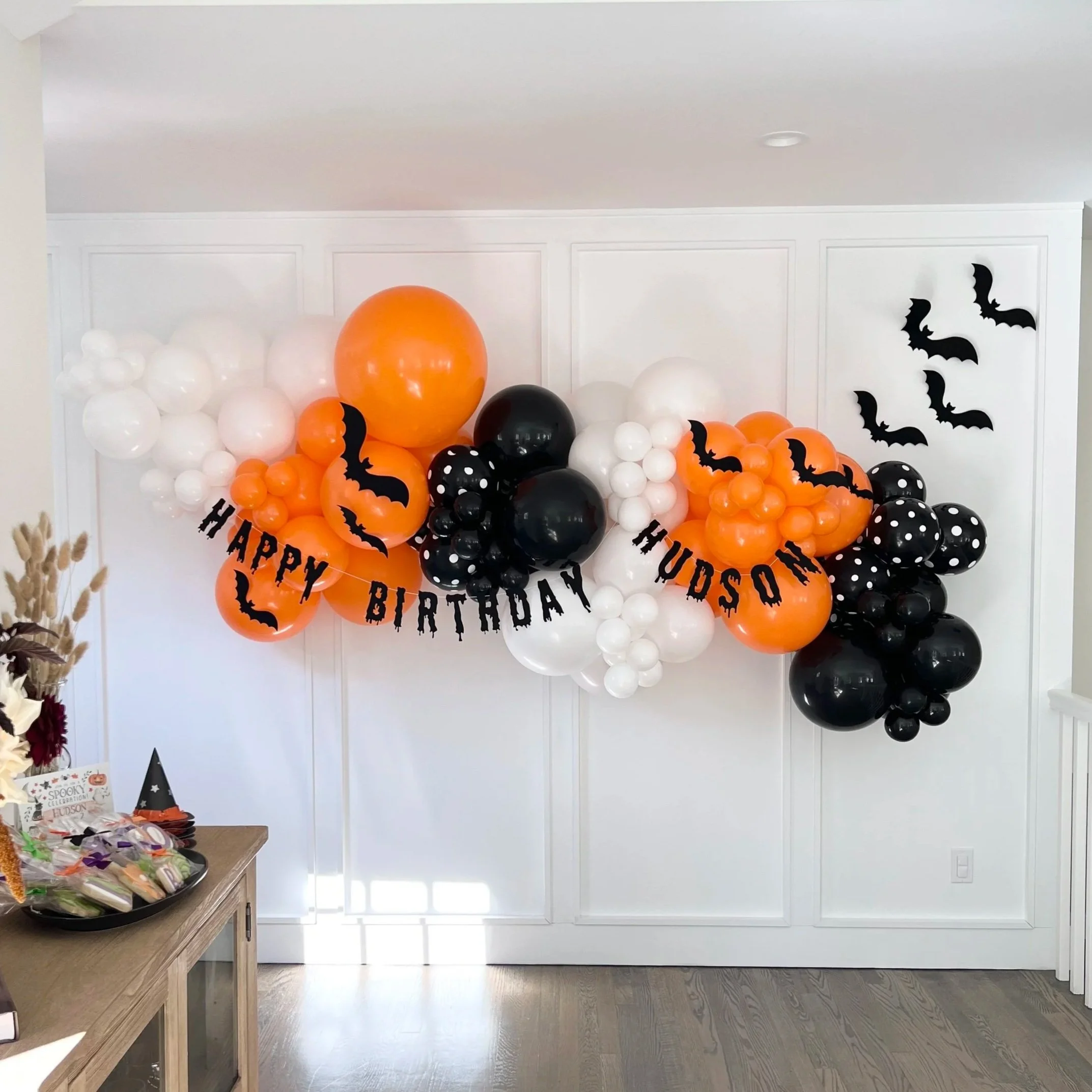 Halloween-themed birthday balloon decoration with orange, black, and white balloons, featuring bat cutouts and a "Happy Birthday Hudson" banner in black letters, on a white wall.