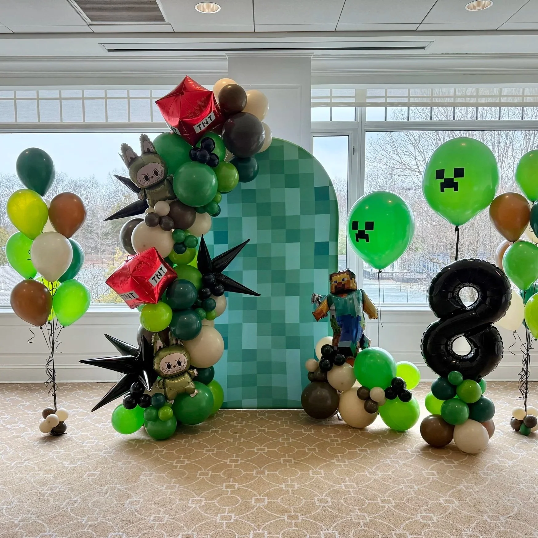 Minecraft-themed balloon decoration with pixel art backdrop, featuring character balloons, Creeper face balloons, TNT block balloons, and a large number 8 balloon, set against a large window with trees outside.