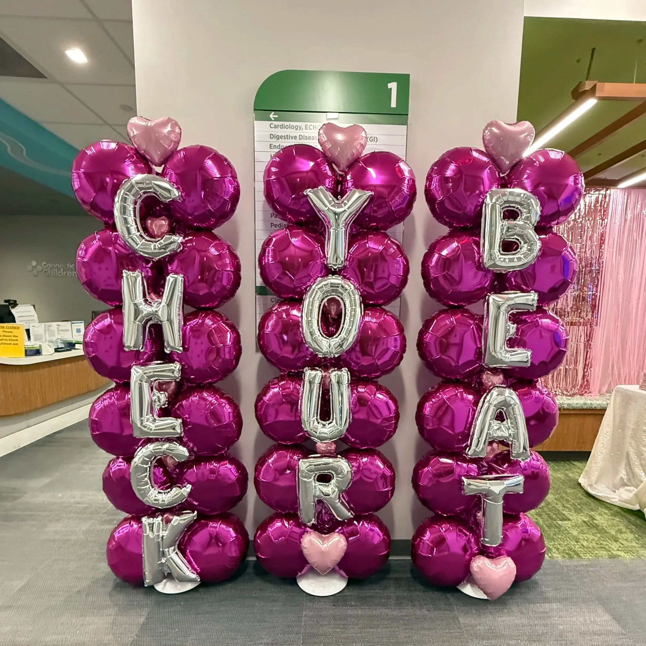 Decorative balloon columns with pink heart-shaped and round balloons spelling out 'CHER' 'YOUR' 'BREAT' with additional pink heart balloons at the bottom, set up in a public space.
