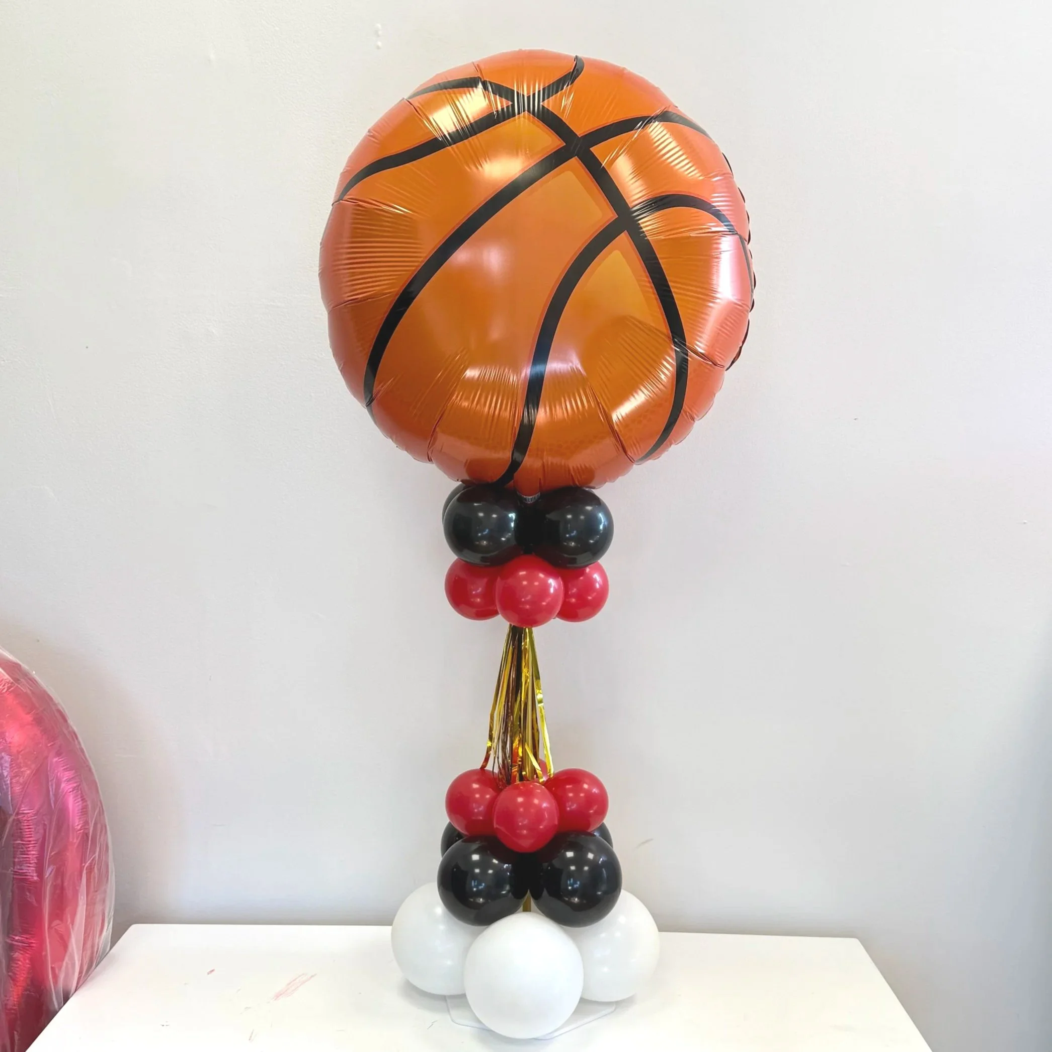 A basketball-shaped balloon attached to a cluster of red, black, and white balloons, with gold curling ribbon at the base, displayed against a plain white wall.