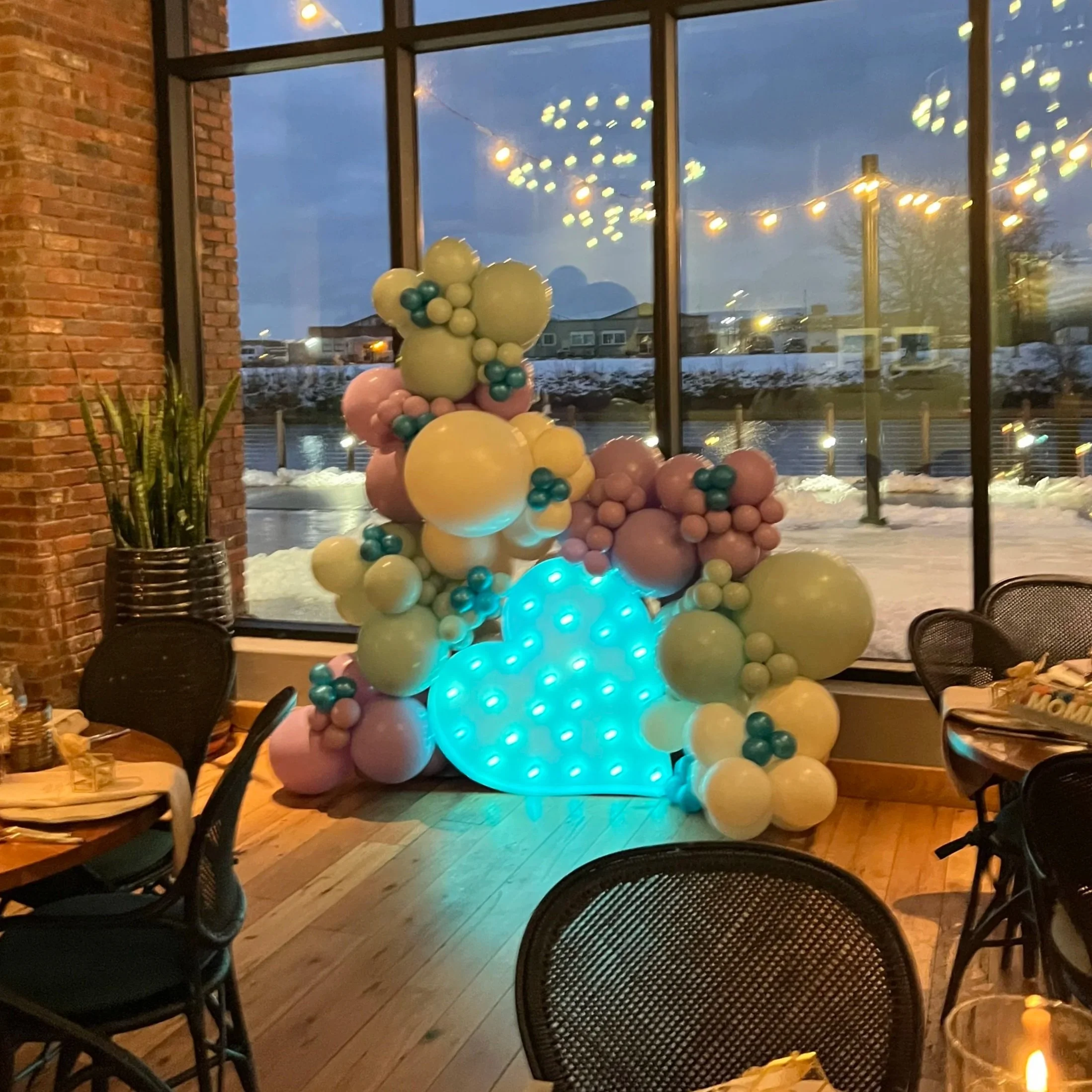 Balloon display with pastel-colored balloons and a lighted cloud shape, set inside a venue with large windows showing snow outside at dusk.