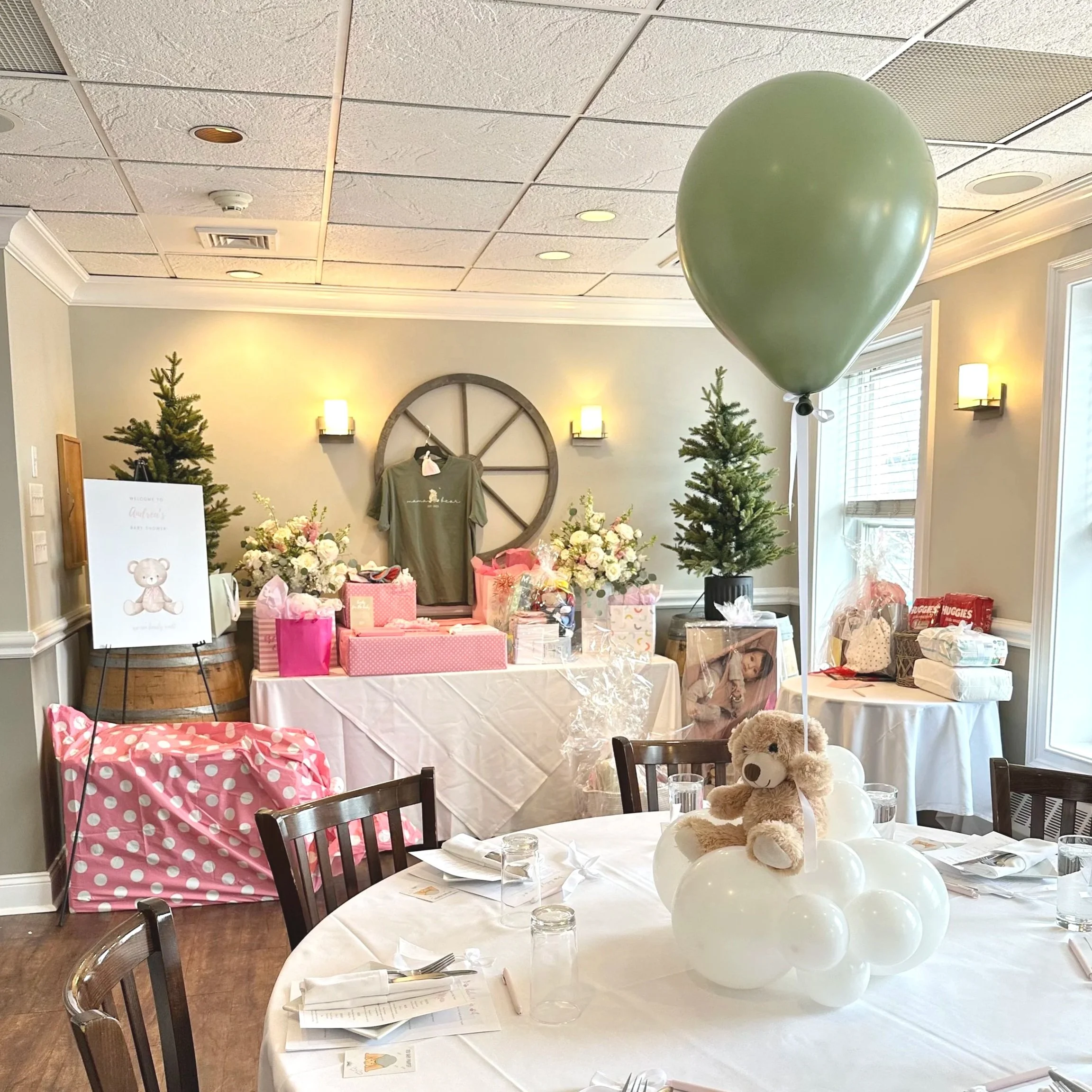 Baby shower setup with a table decorated with a stuffed teddy bear, white balloons, and a cloud-shaped centerpiece with balloons. In the background, a table displays gifts, flowers, and decor, with a large green balloon floating nearby. The room has beige walls, windows with blinds, and wall-mounted lights.