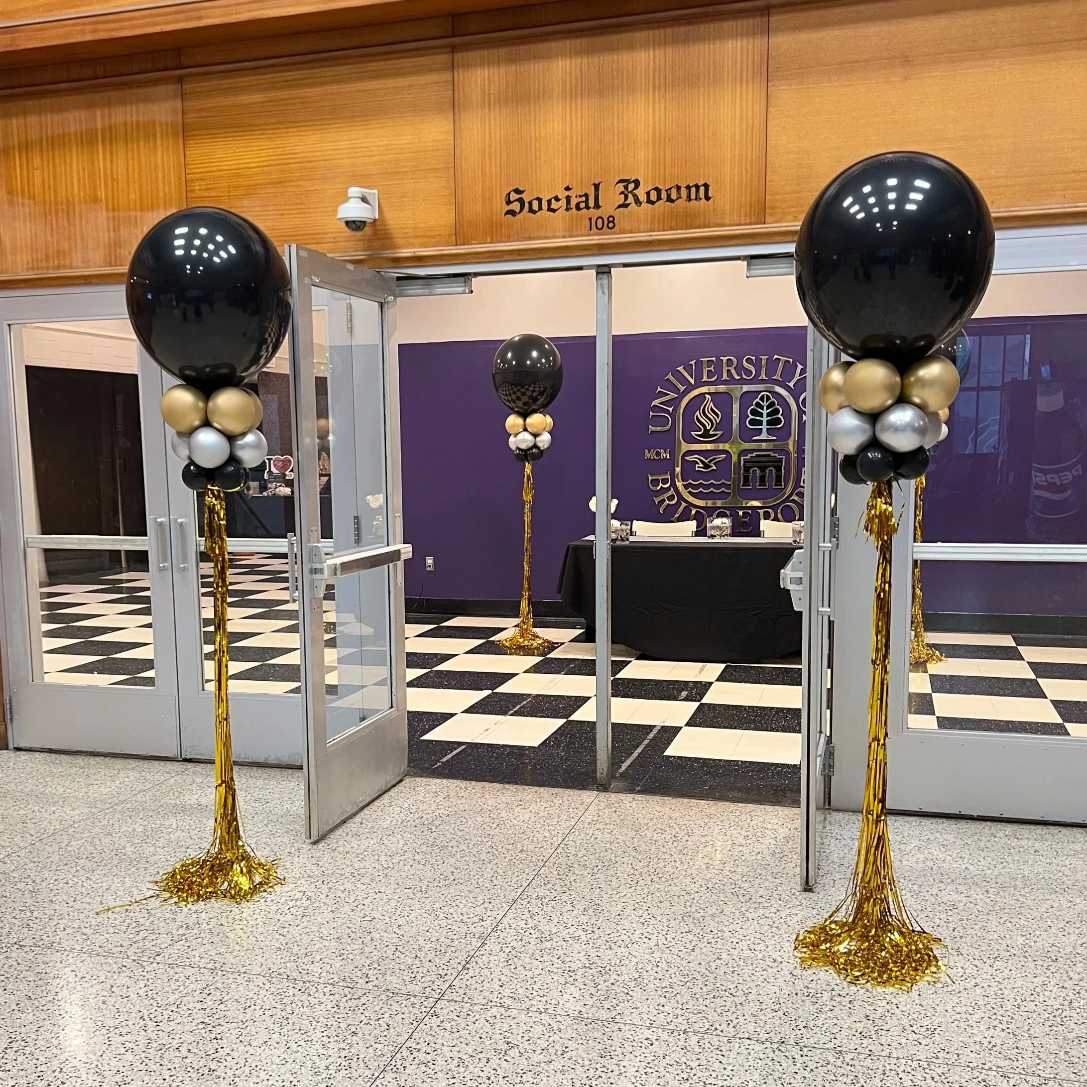 Entrance decorated with black, gold, white, and silver balloons with gold tinsel at the base, leading into a room with a purple wall and a university logo in the background.
