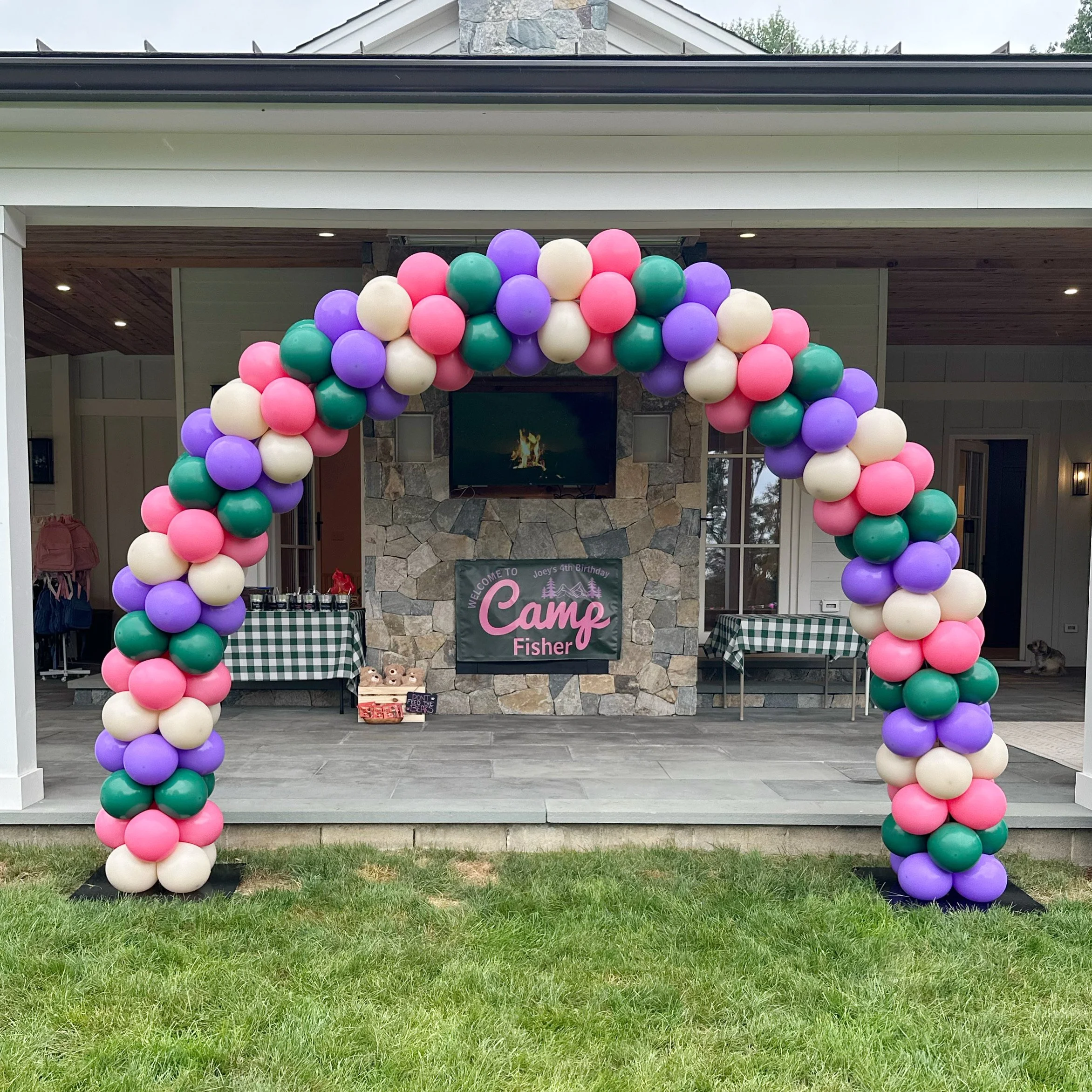 Colorful balloon arch with pink, purple, green, and white balloons at an outdoor birthday party, with a stone fireplace and a banner that reads "Camp Fisher" in the background.