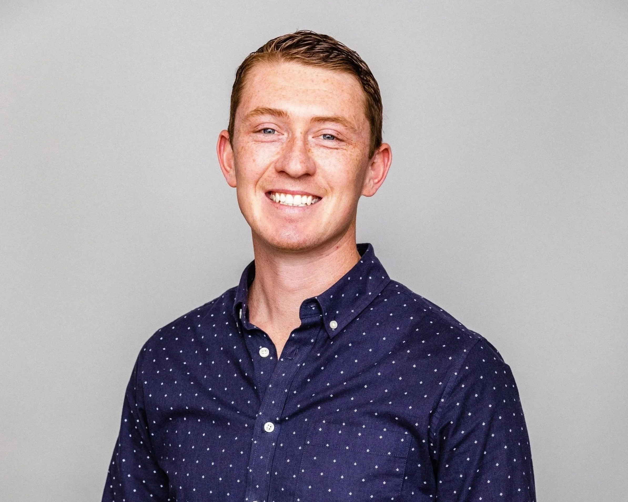 A smiling young man with short red hair and freckles, wearing a navy blue shirt with white polka dots, standing against a plain gray background.