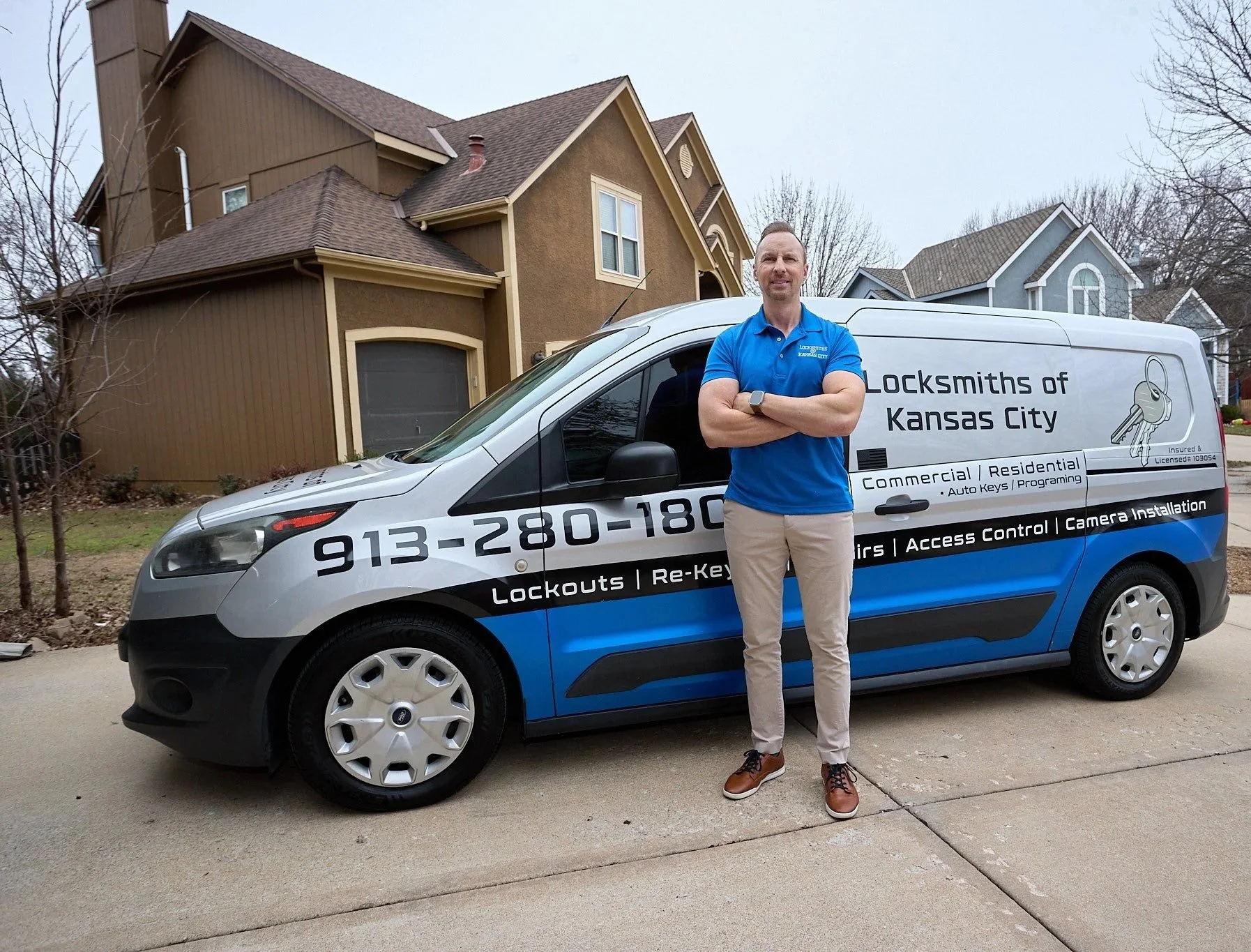 A man standing in front of a locksmith service van with the text 'Locksmiths of Kansas City' and a phone number, parked on a residential street with houses in the background.